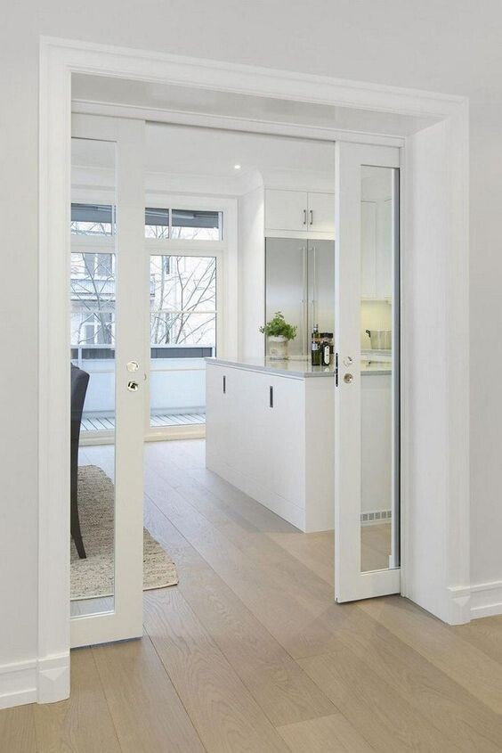 Open white glass doors lead into a bright kitchen with island, window fronts and wooden floor.