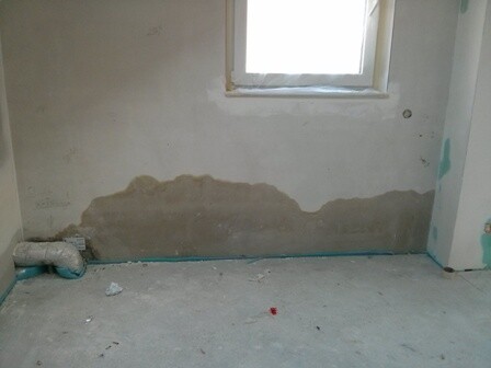 Unbearbeiteter Innenraum mit feuchter Wand, kleinem Fenster und links liegendem Rohr