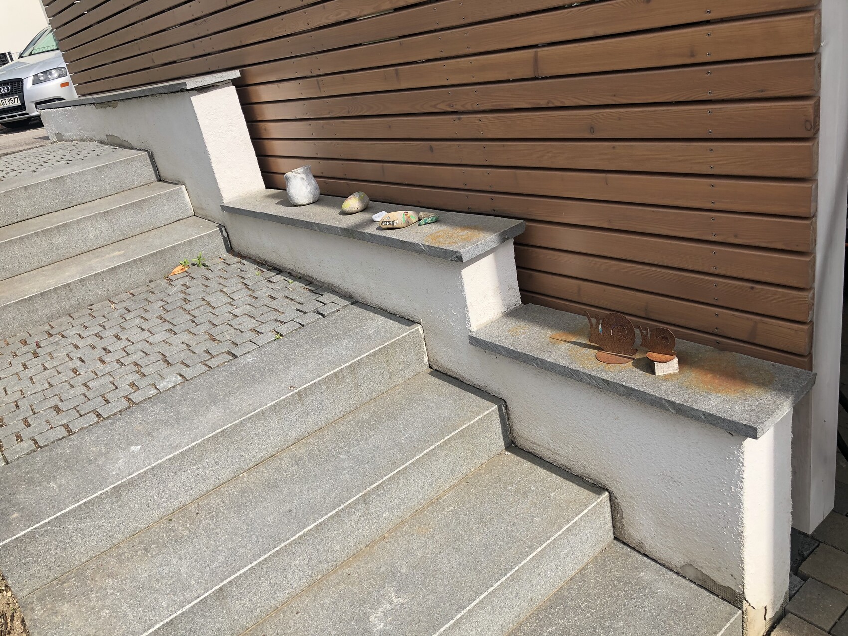 Exterior concrete steps, stone bench with stones and rusty metal figures; car