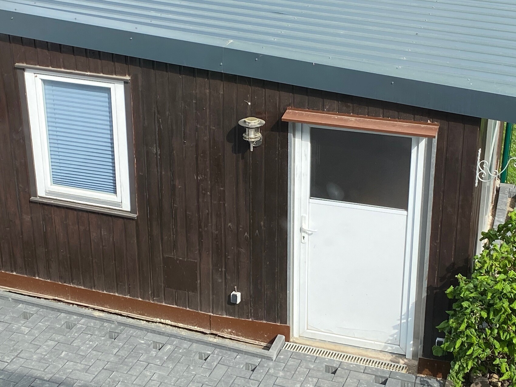 Holzfassade eines Gebäudes mit weißer Tür, Fenster mit Rollladen, Außenleuchte, Busch rechts.