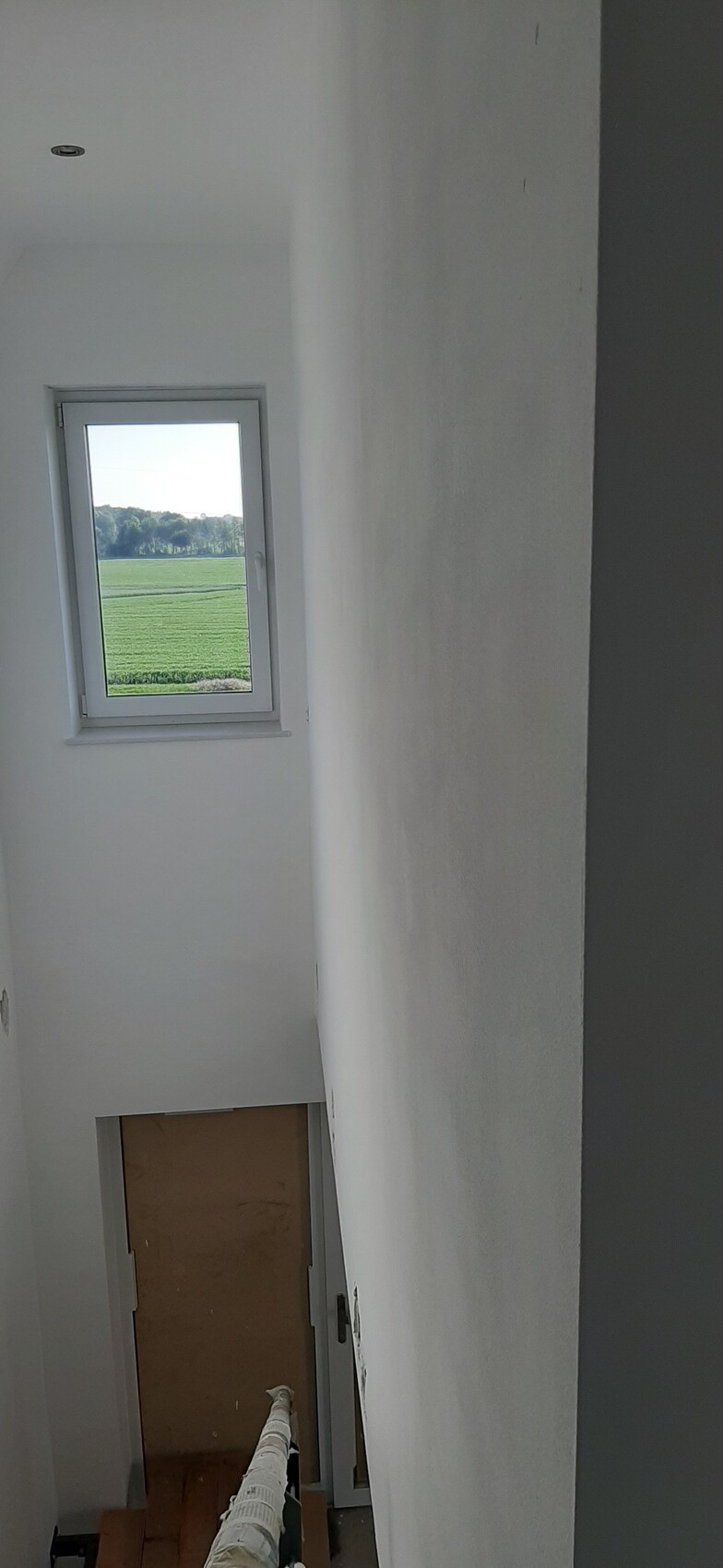 Heller Treppenaufgang mit kleinem Fenster, Blick auf grüne Felder außerhalb; weiße Wände.