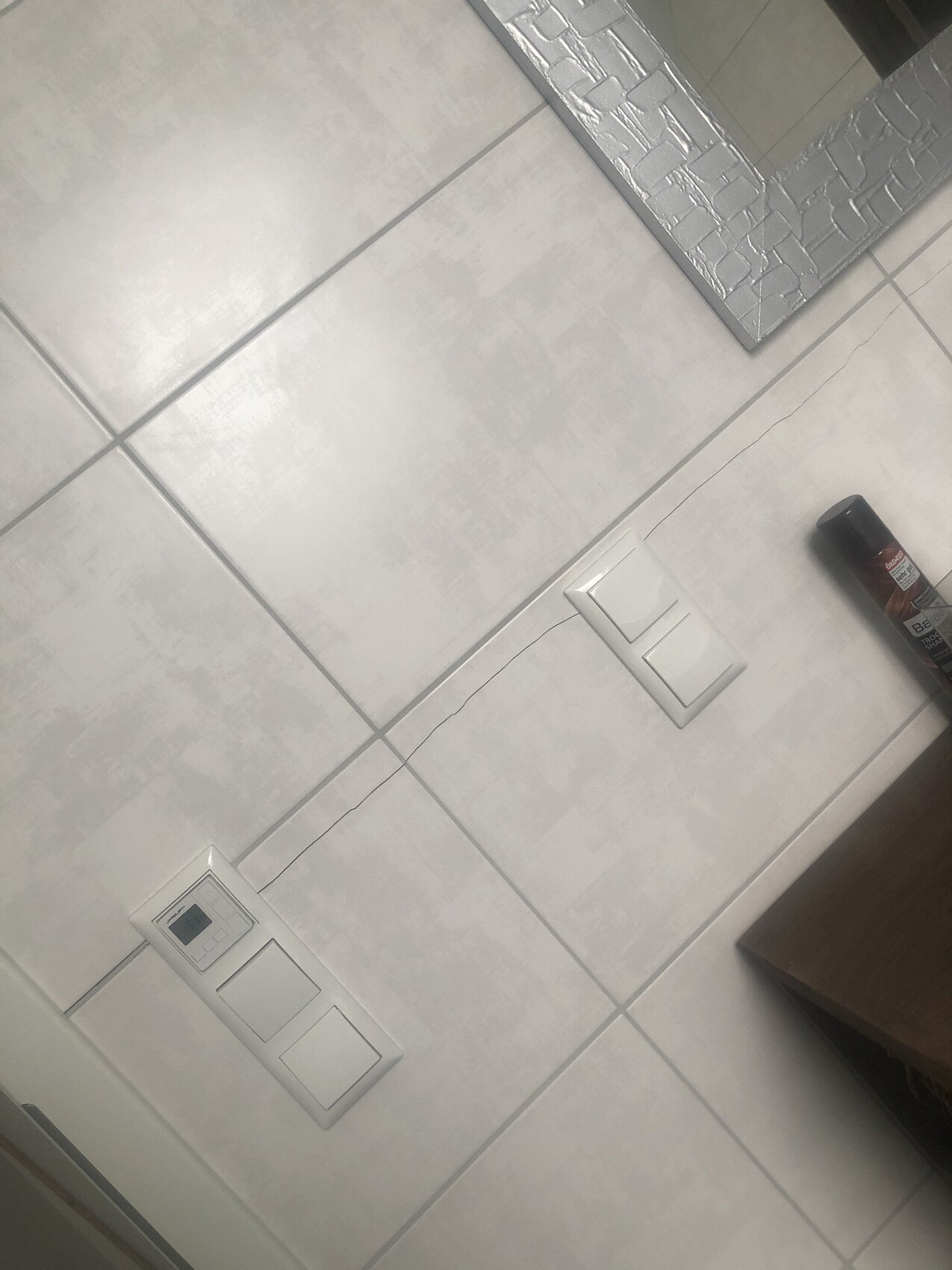 Grey tiled wall with two switches, thermostat on the left, mirror with mosaic frame; spray can on the right.
