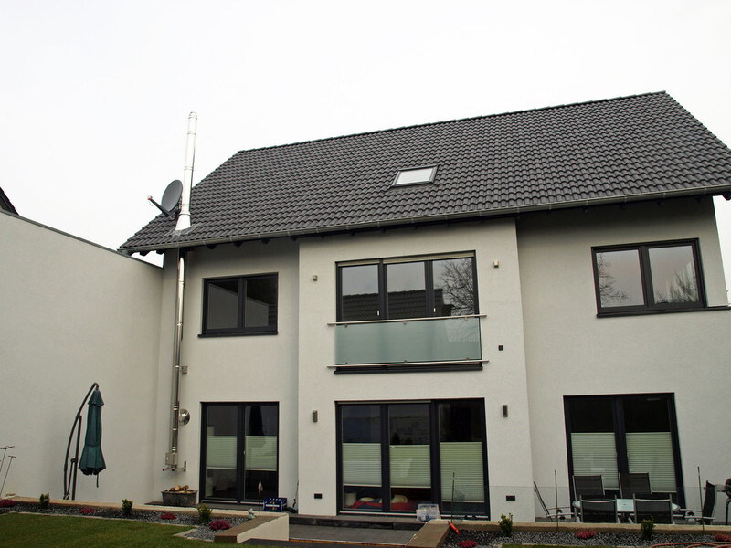 Modern two-story house with dark tiled roof, glass balcony, large windows and garden.