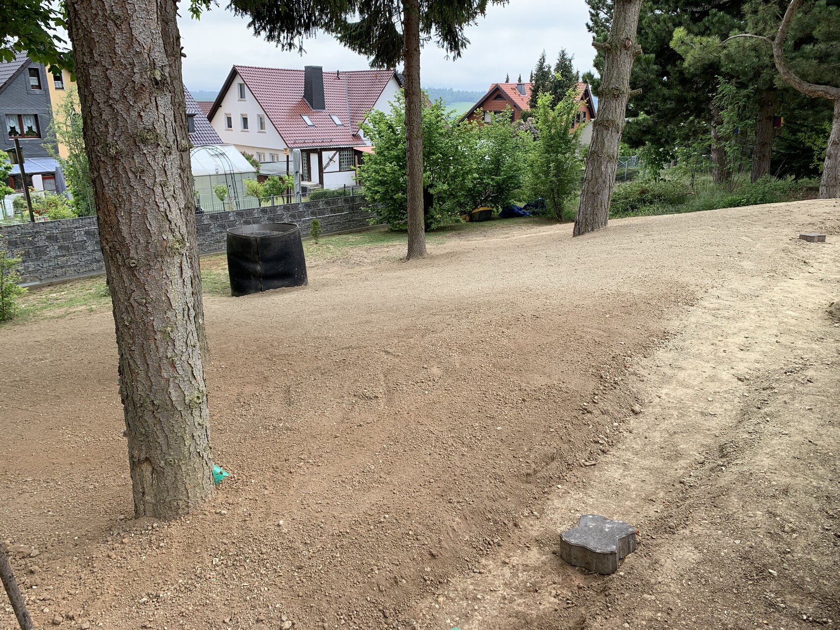 Parkfläche mit Kies- und Sandboden, mehrere Bäume, Häuser im Hintergrund und ein schwarzer Behälter.