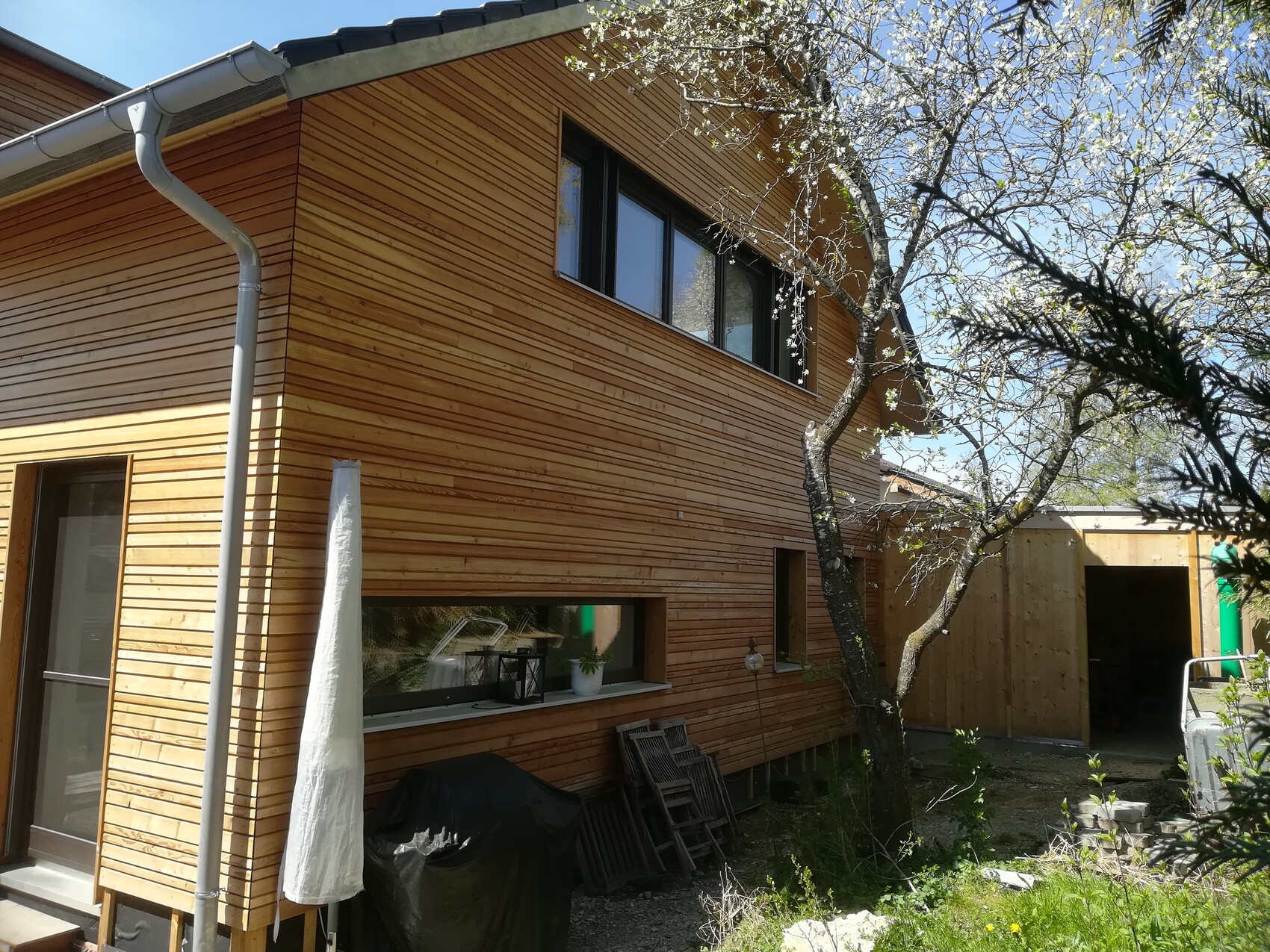 Wooden house with horizontal cladding, flowering tree in front, shed on the right.