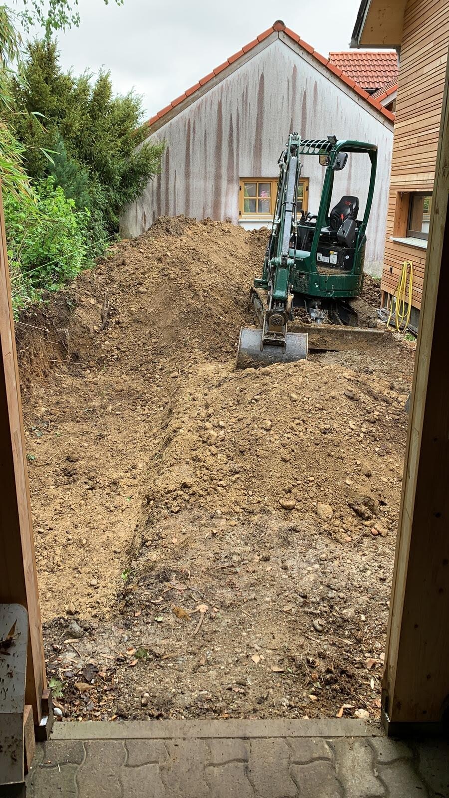Small excavator digging foundation pit next to residential house on a construction site
