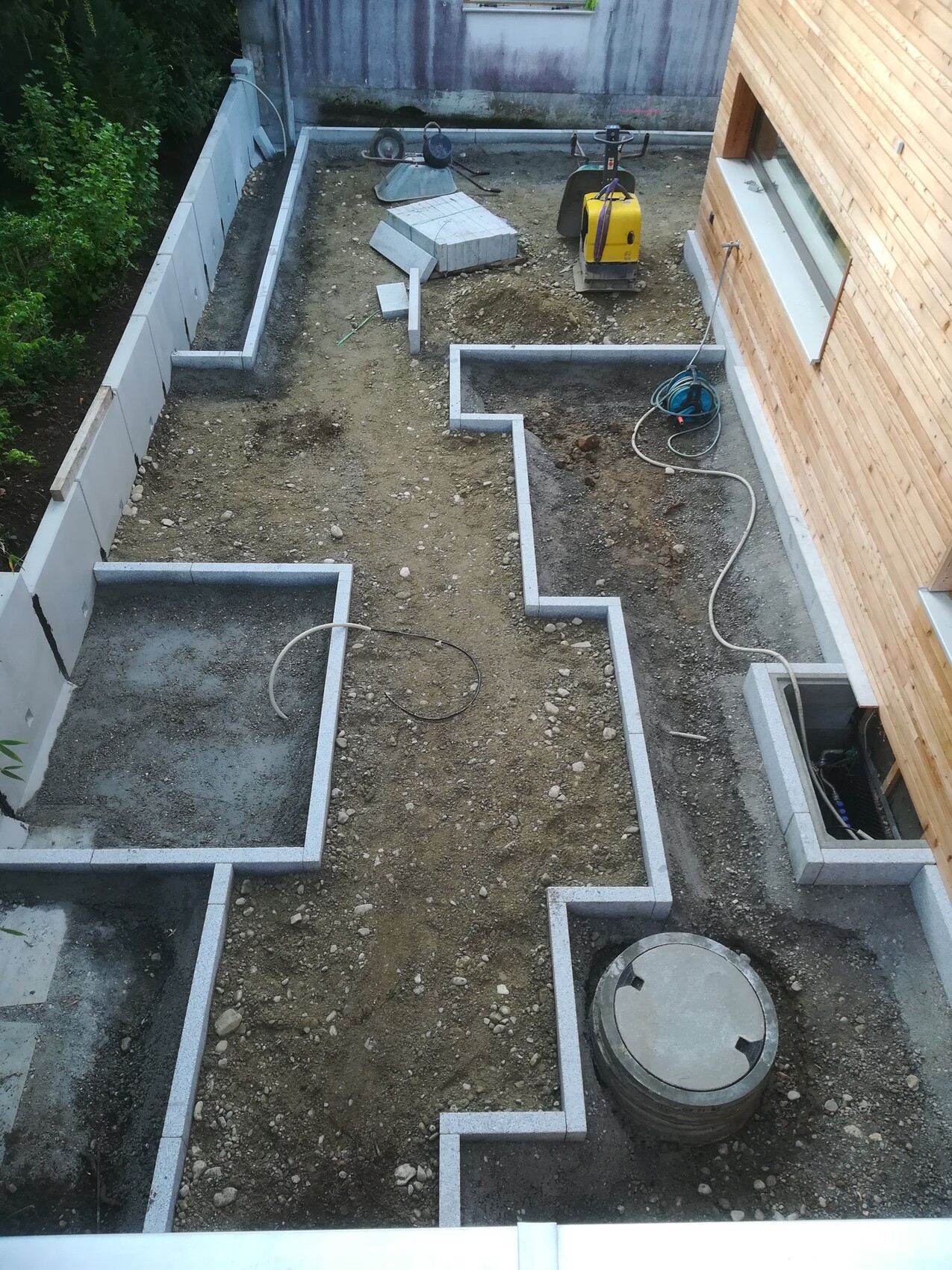 Construction site: layout of concrete foundations on gravel, yellow plate compactor, hose, round cover.