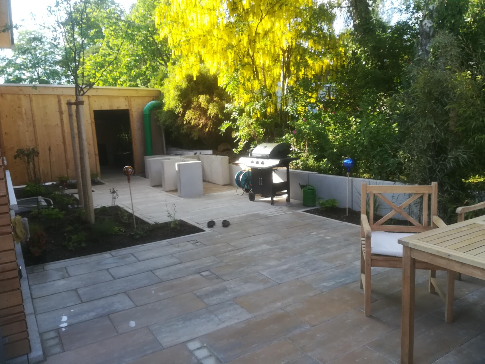 Garden terrace with wooden table and chairs, grill, planting beds and green hedge in the background.