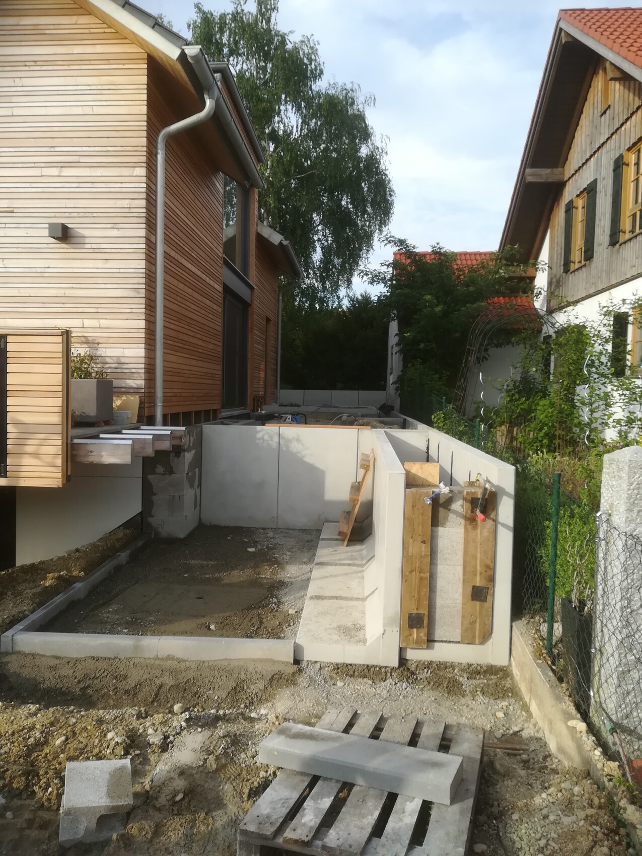 Construction site between wooden houses: open concrete foundation walls, construction timber, gravel and pallets.
