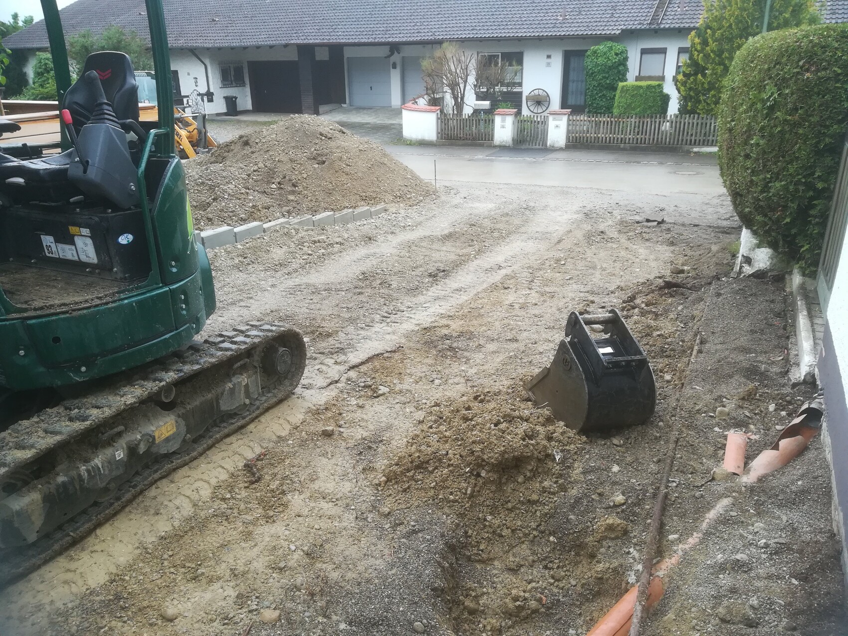 Green tracked excavator at roadside, soil piles, shovel next to excavation pit, houses in the background.