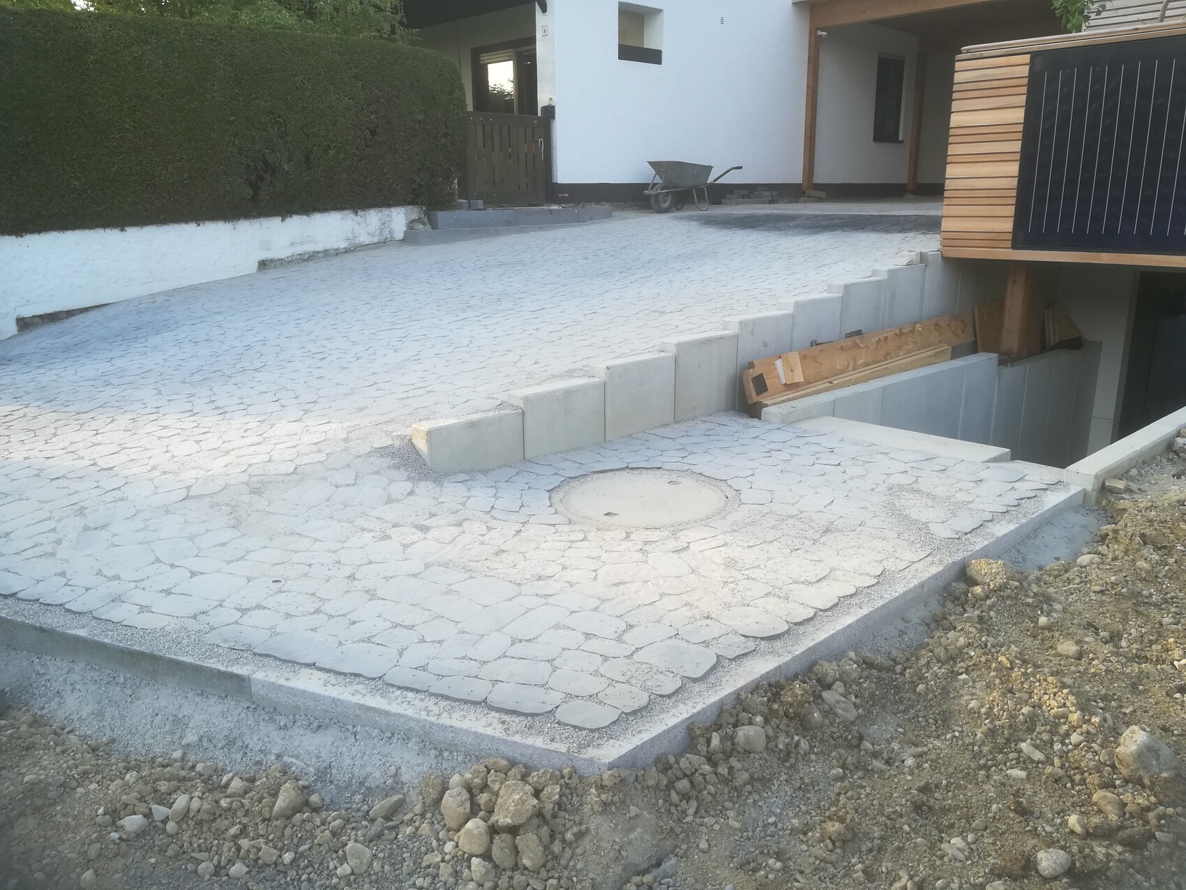 Paved driveway in front of house under construction; round manhole cover, wooden slats, wheelbarrow in background.