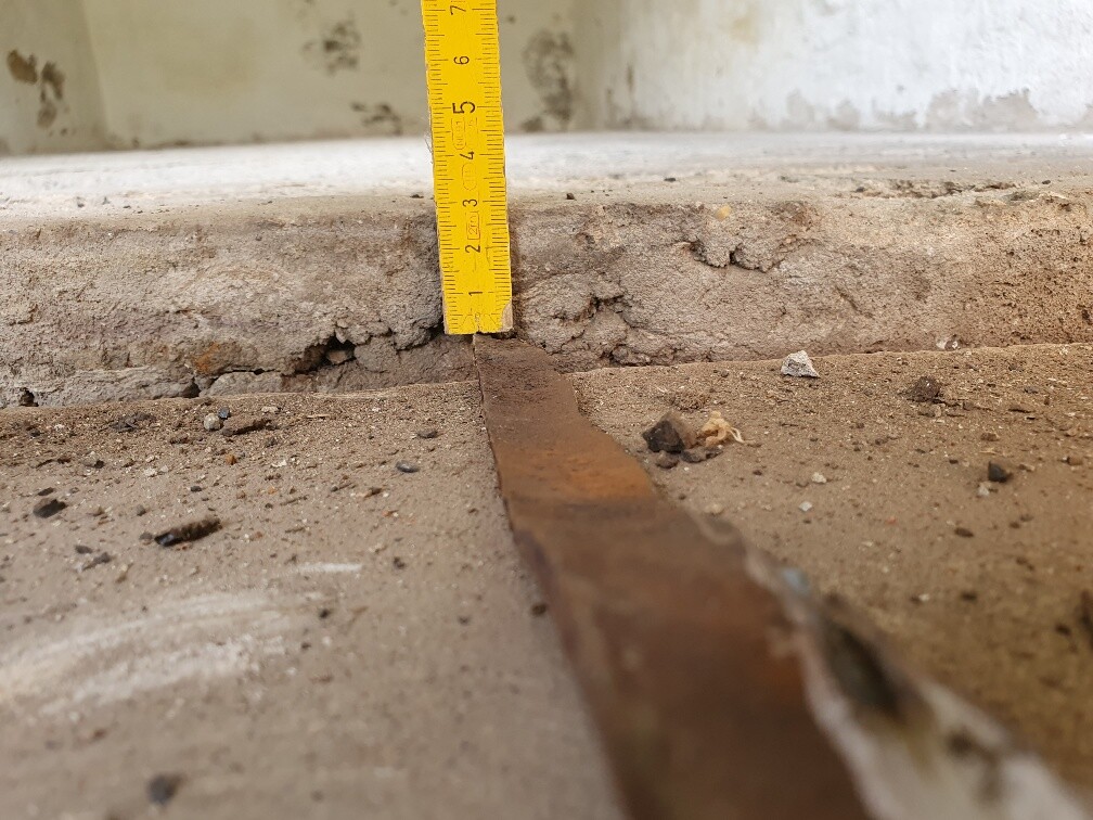 Yellow measuring tape standing vertically against rough concrete edge, rusty metal strip lying on the floor.