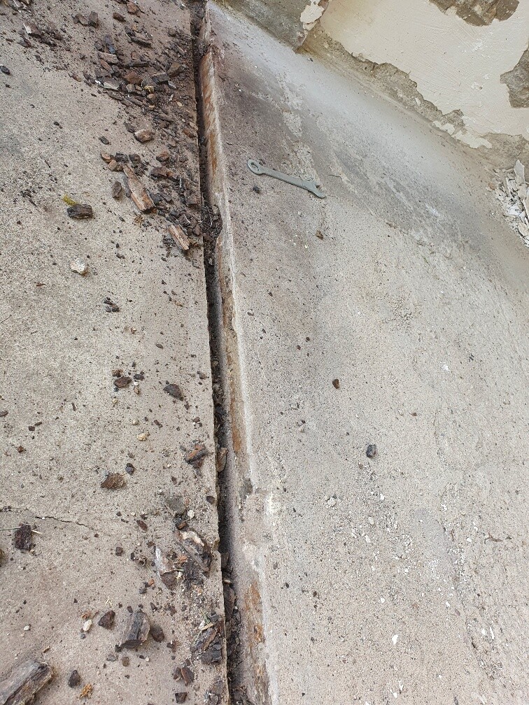 Deep gap in a concrete slab, surrounded by rubble and plaster remains; small metal piece lying on top.