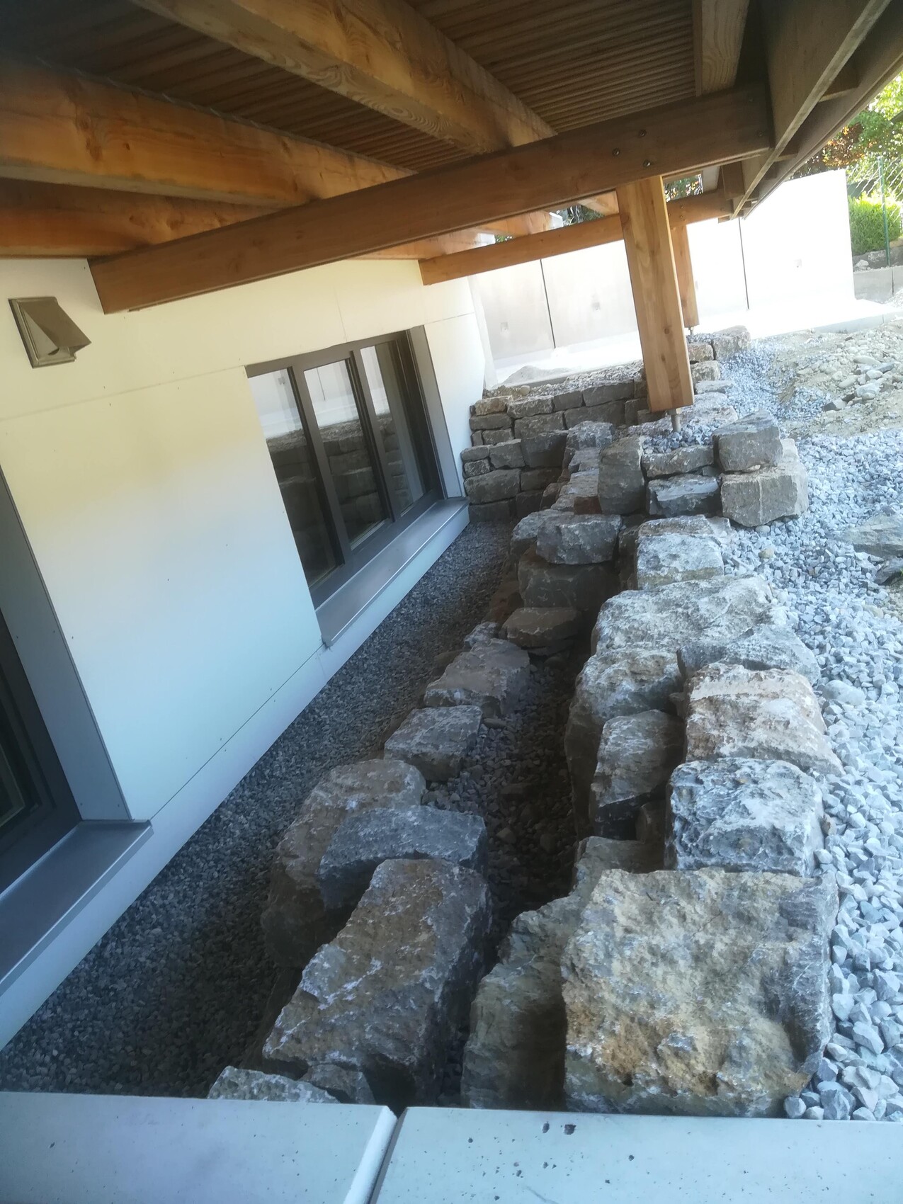 Outdoor area: Stone wall as retaining wall along the house, under deck beams, gravel ground.