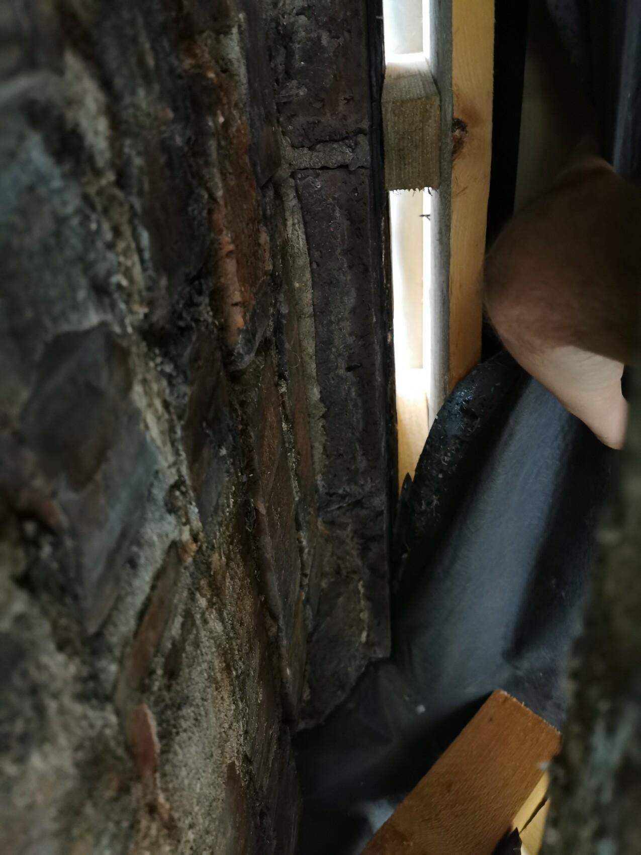Narrow gap between brick wall and wooden frame, visible light gap; hand and dark cloth in front