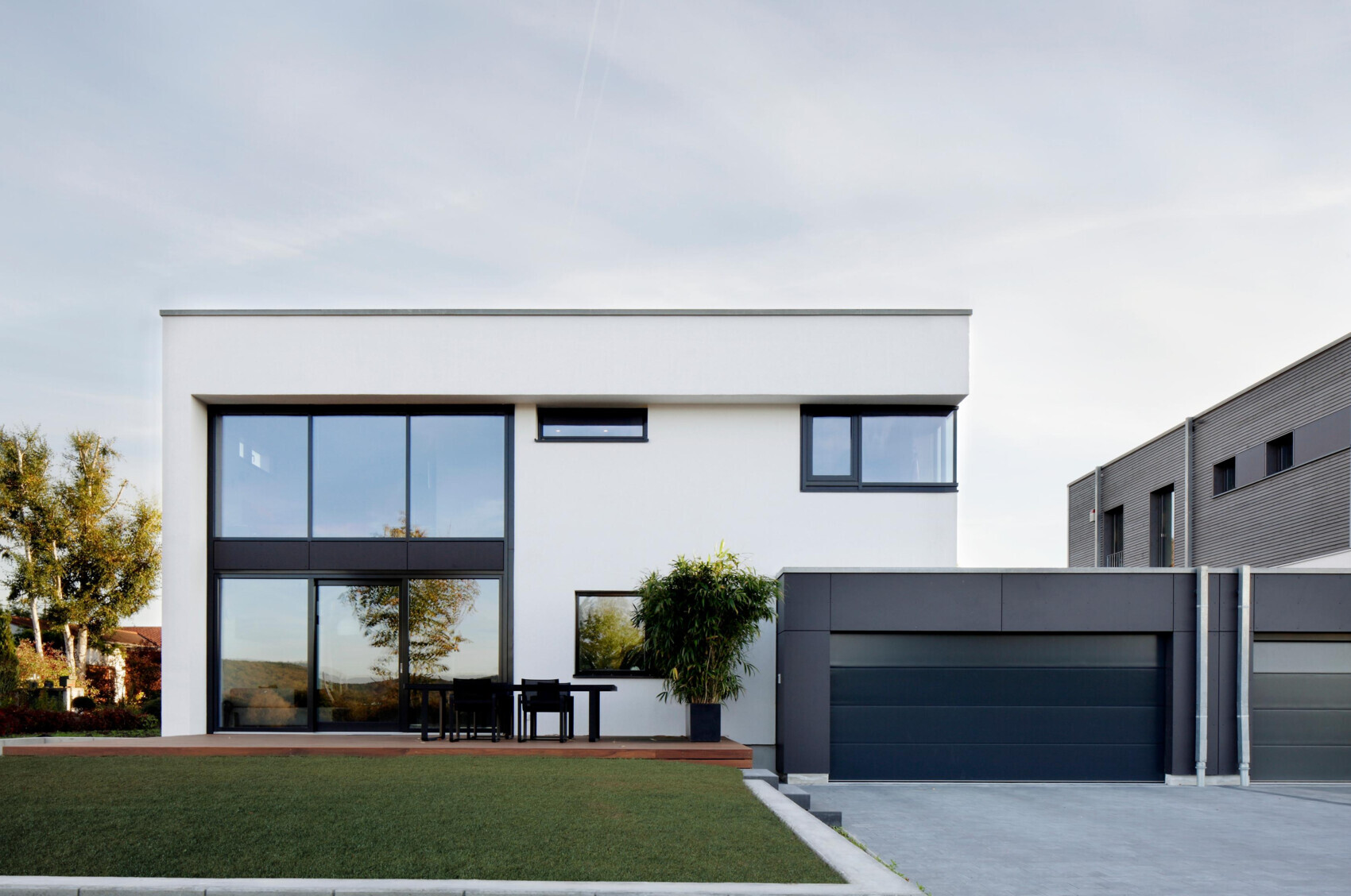 Modern white villa with large glass facades, terrace with table and chairs, dark garage on the right.