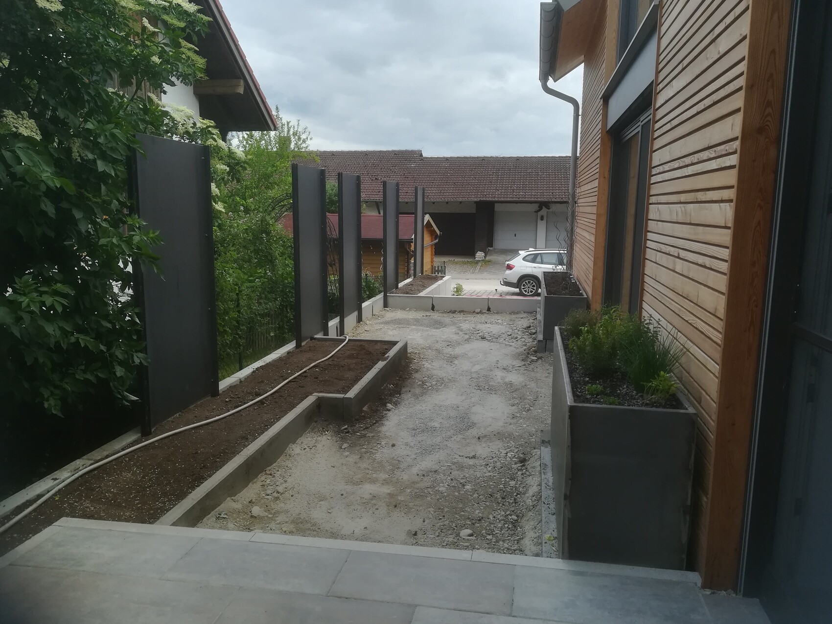 Path between wooden house on the right and hedge on the left; dark posts, planter boxes, car in the background.