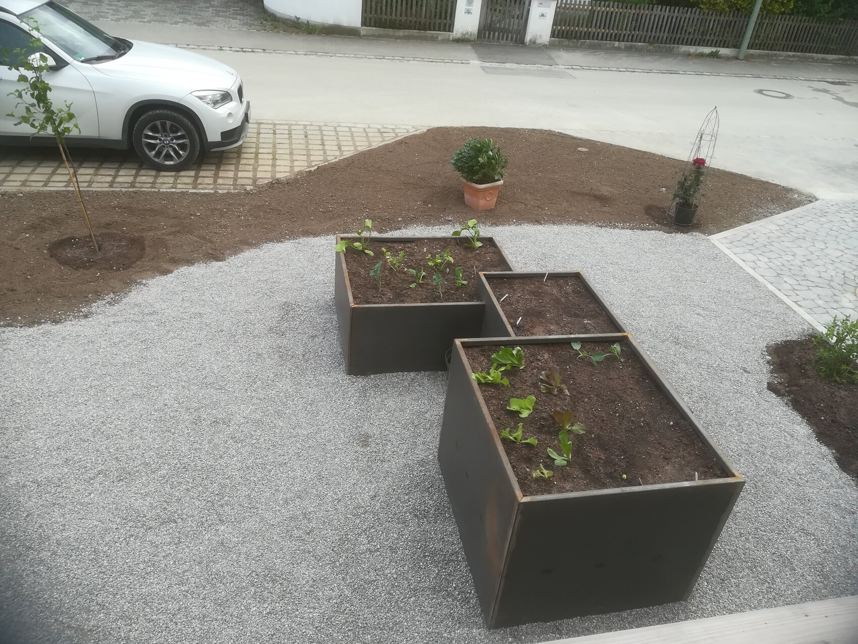 White car parked next to rectangular metal planter boxes with soil and young plants on gravel path.