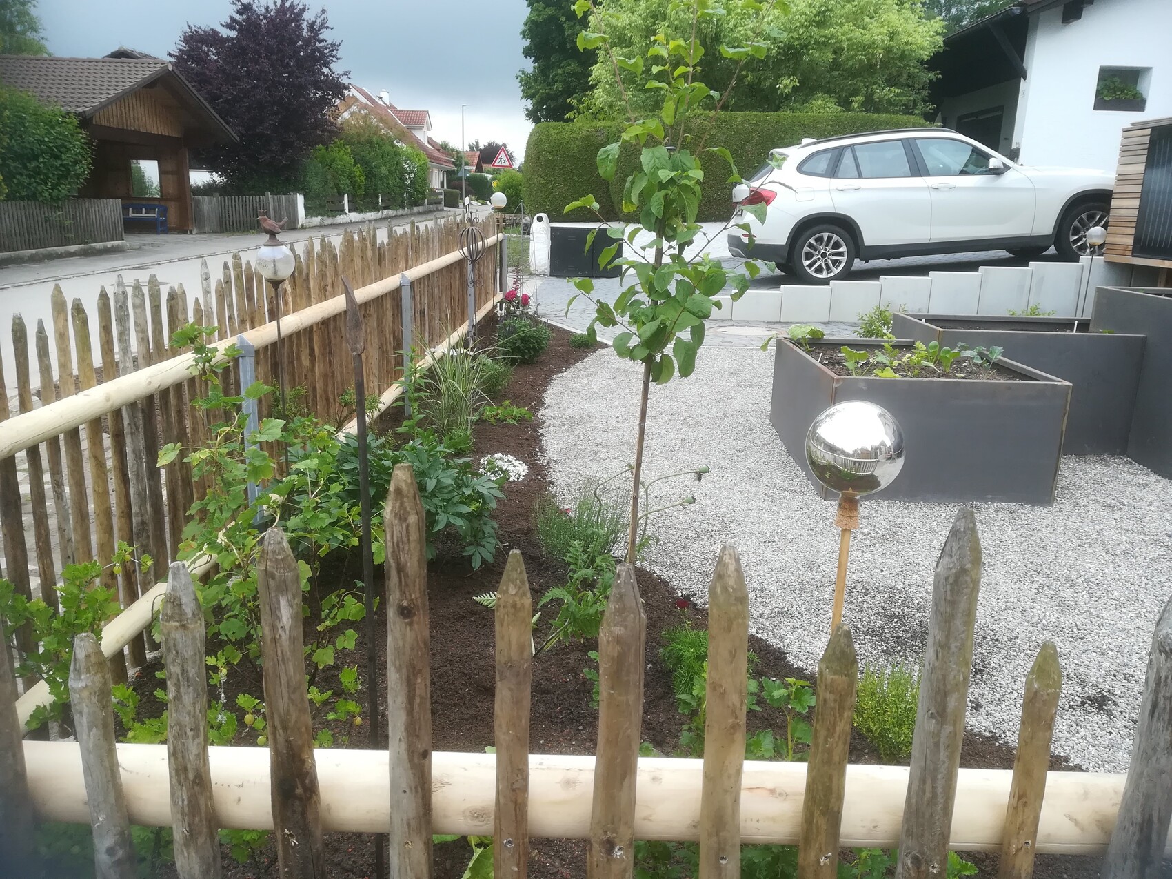 Garden path behind wooden fence with flower beds, tree, globe lamp, and white SUV in the background.