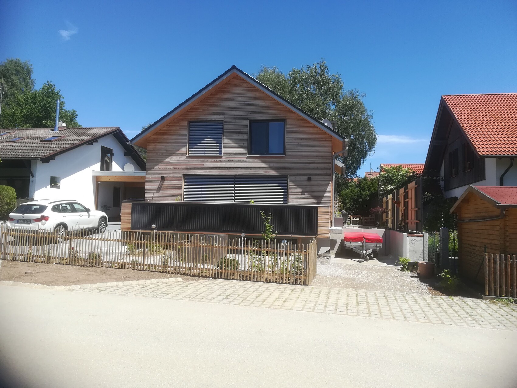 Wooden house in suburb with wood cladding, car on the left, boat trailer with red cover on the right.