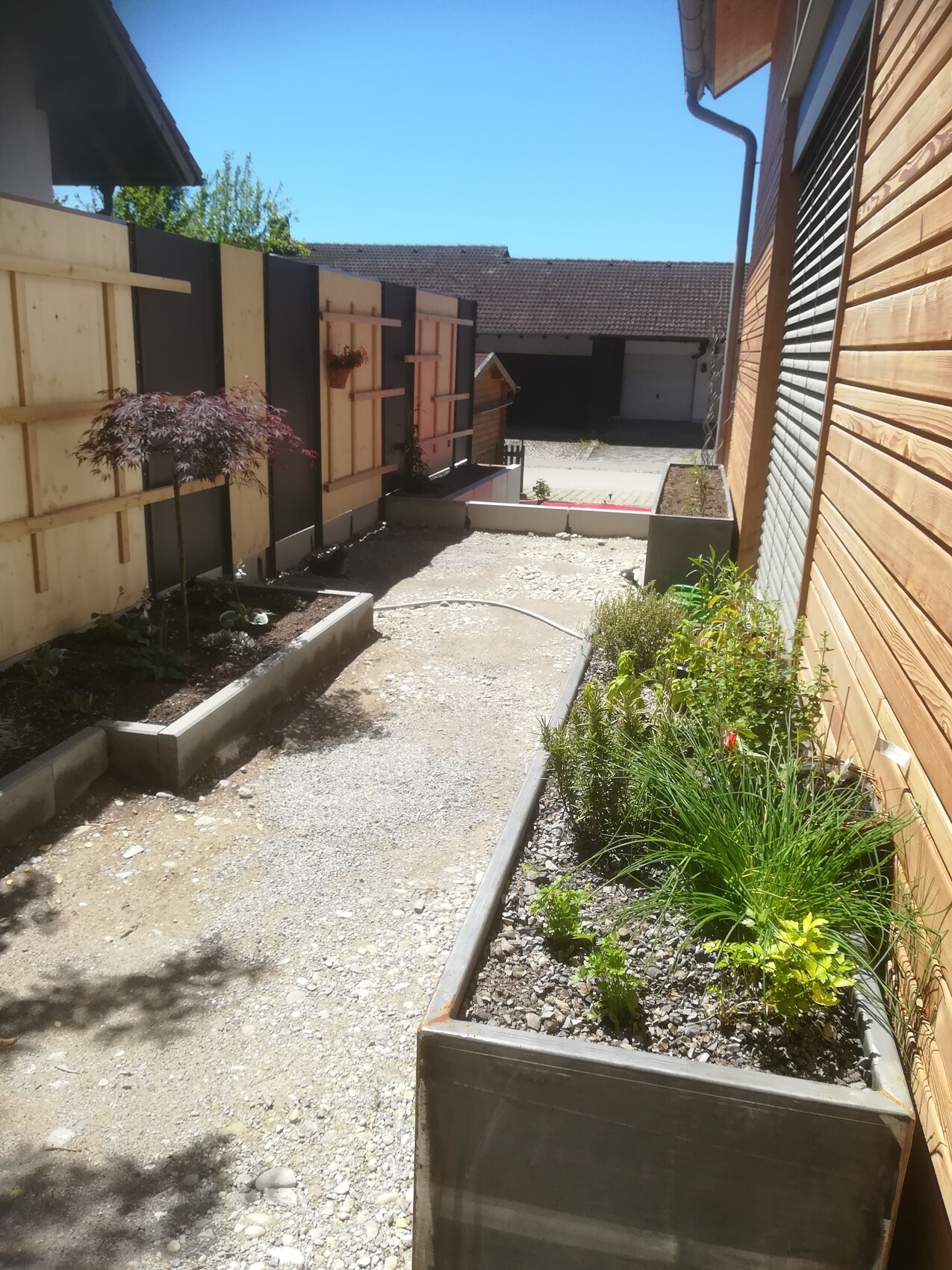 Gravel path between wooden walls (left) and wooden house facade, with planter boxes.
