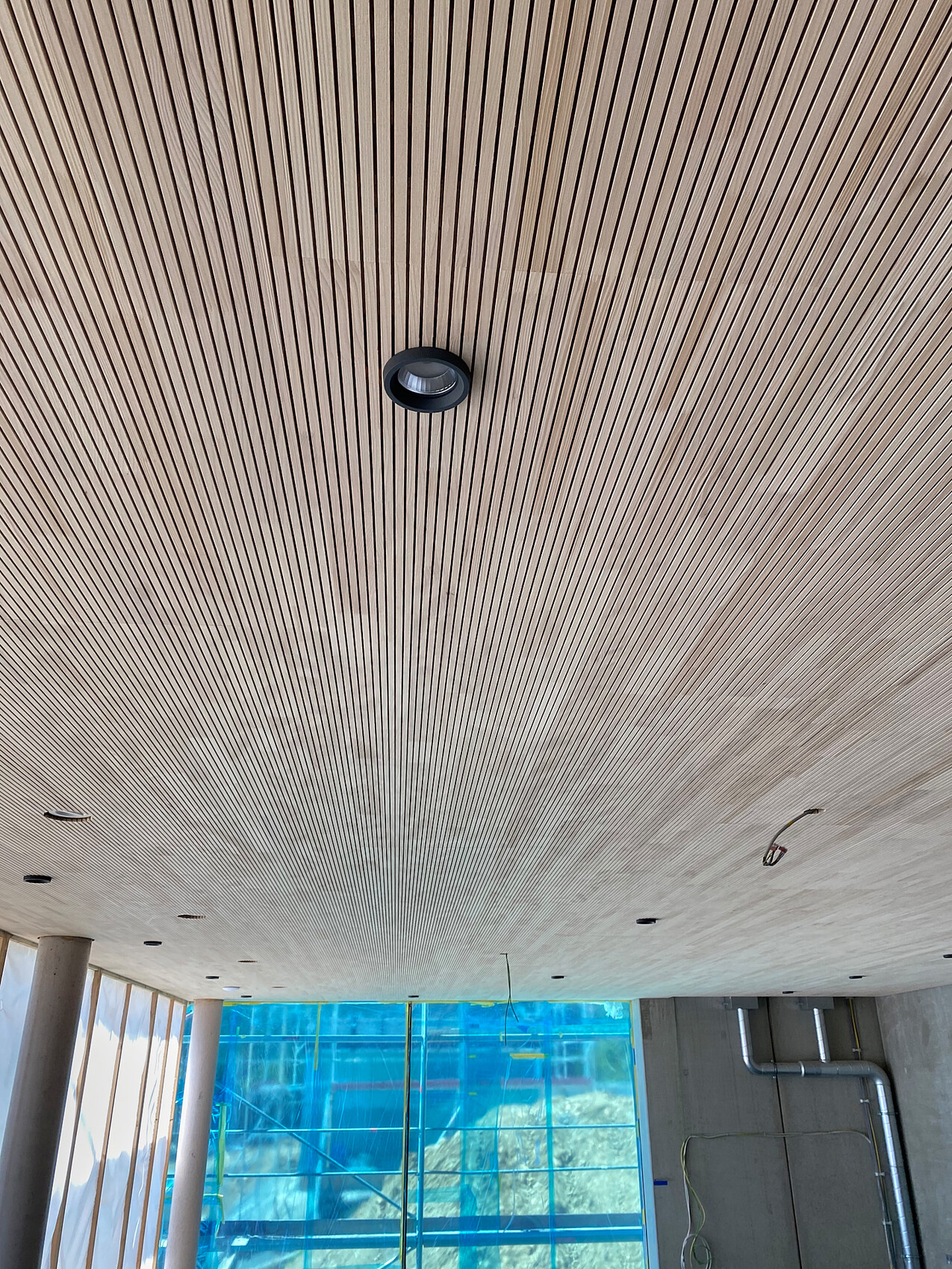 Ceiling with parallel wooden slats; round recessed light, construction site visible behind glass.