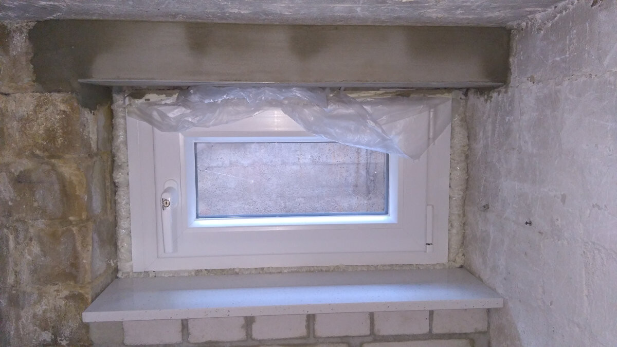 Small white basement window in stone and plaster wall, plastic sheet covering it.
