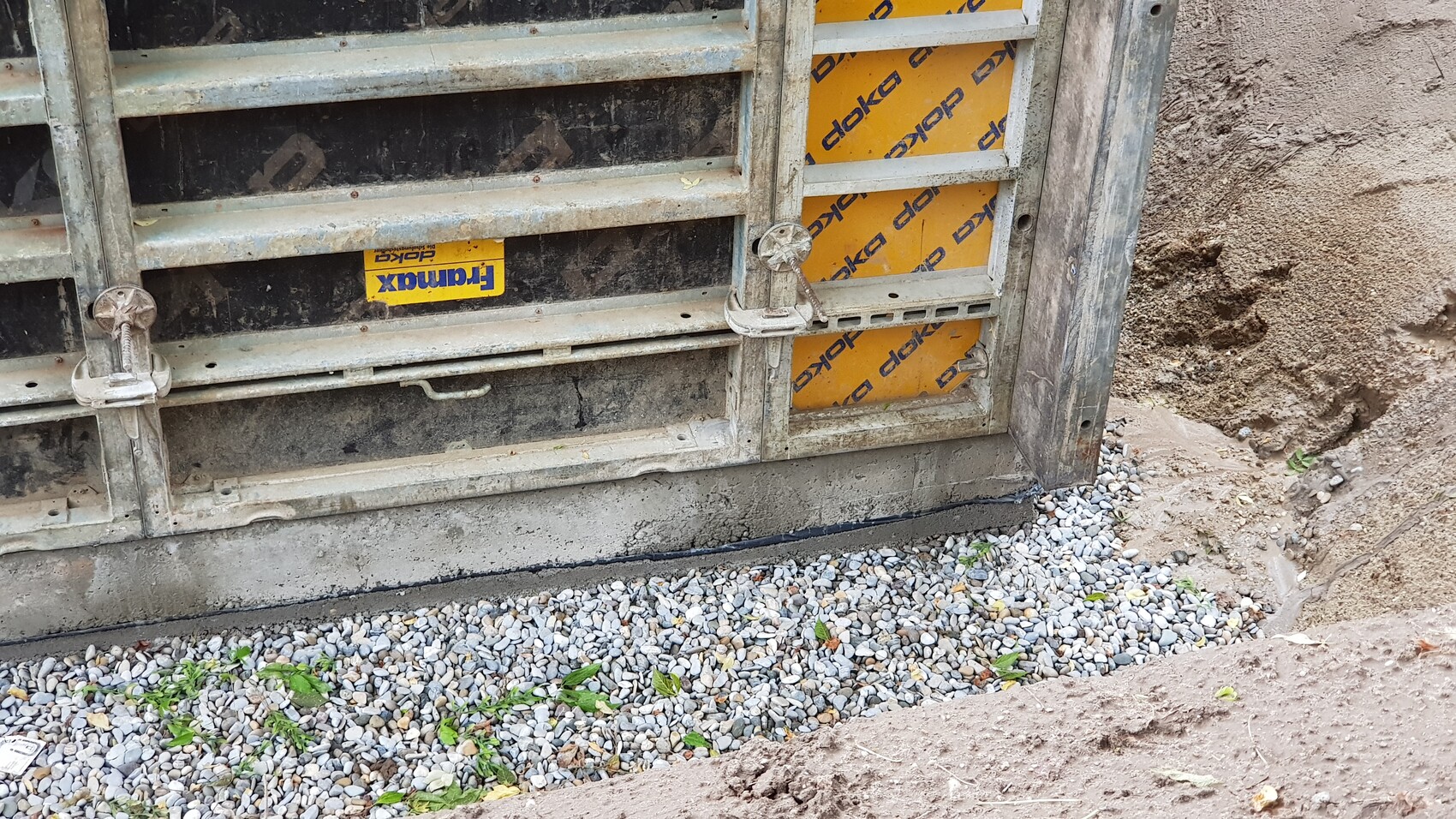 Metal gate with locks at a construction site; yellow formwork panels, gravel and soil.