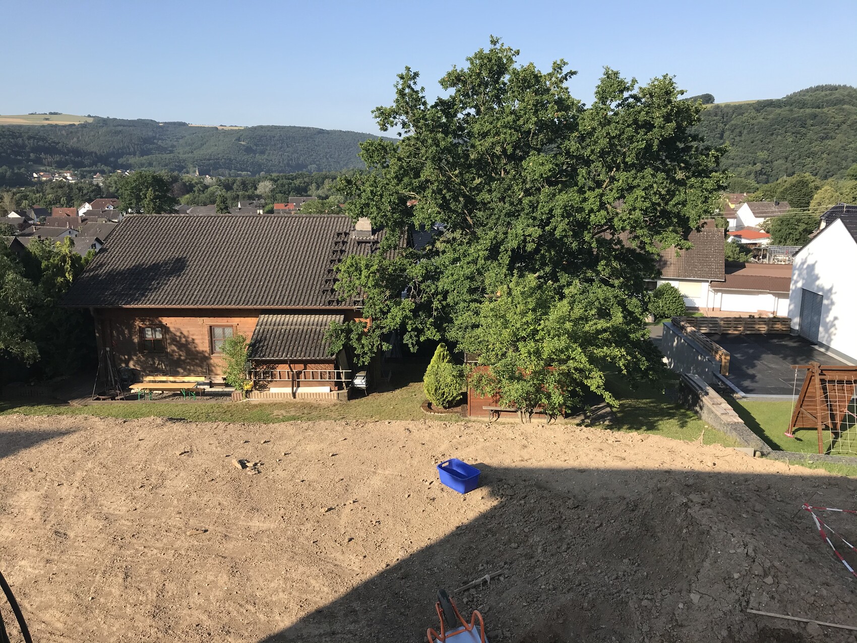 Blick auf ein kleines Haus mit großem Baum im Garten und Baugrund davor.