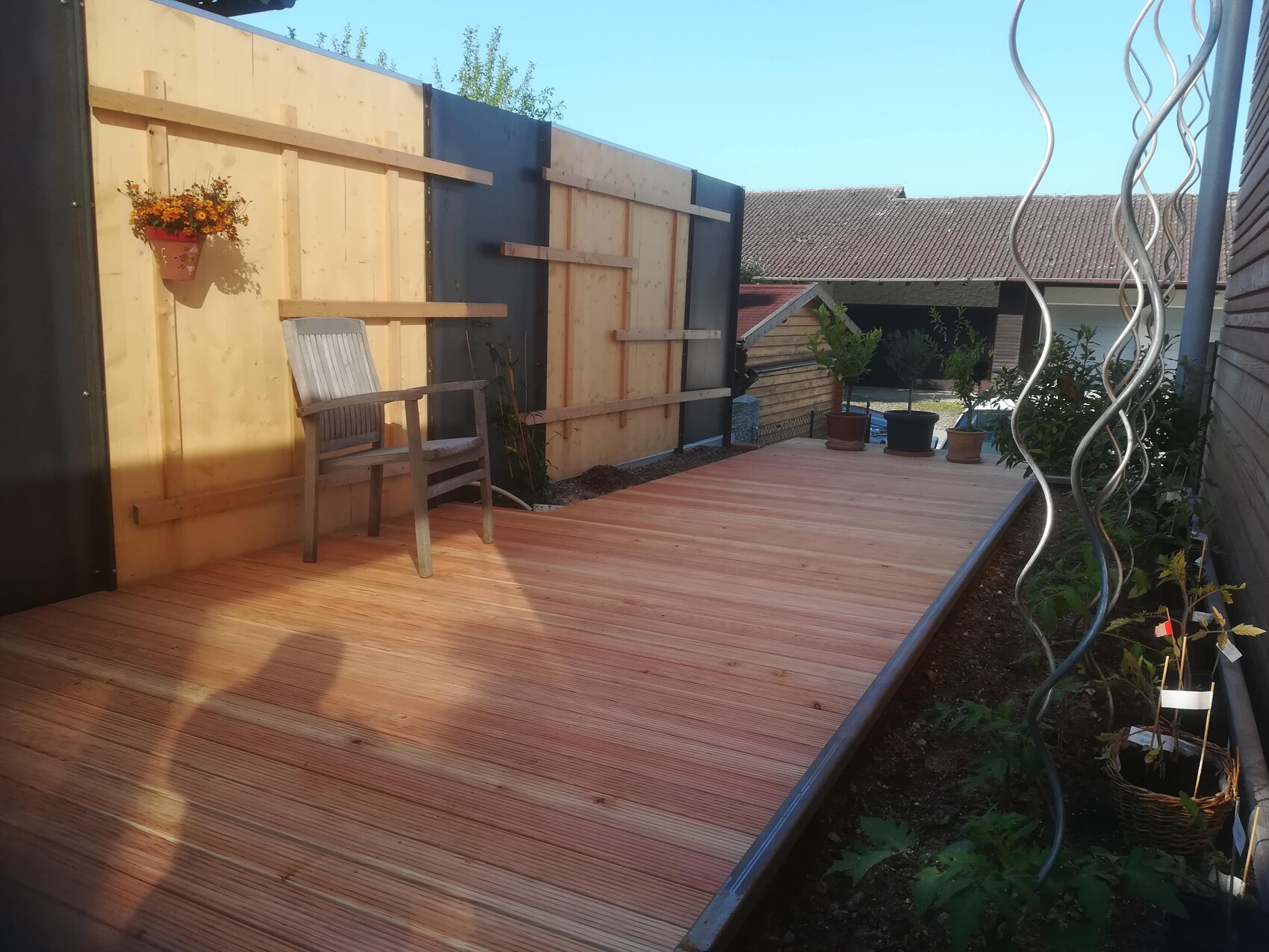 Wooden deck in the garden with gray chair, wooden fence wall, and potted plants; spiral sculpture on the right.