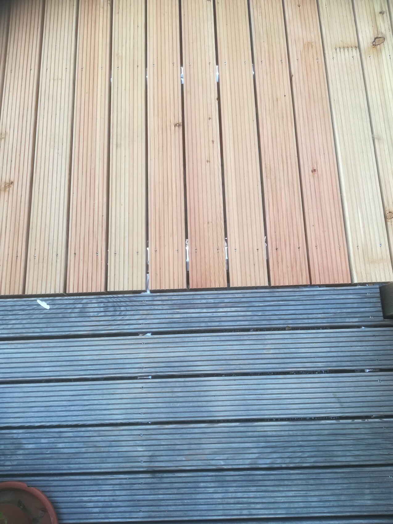 Wooden deck with vertical boards on top; below, gray grooved planks, left planter visible.