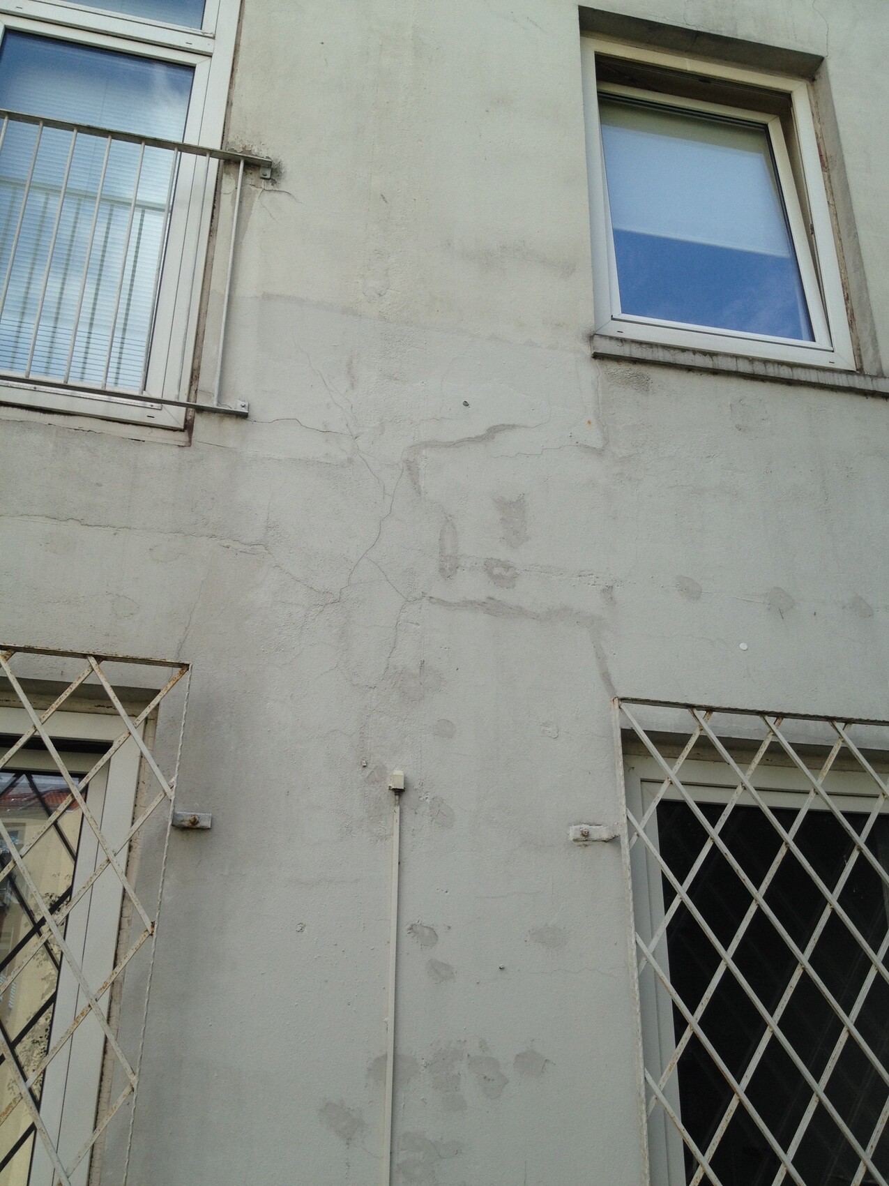 Außenwand eines Gebäudes mit Rissen; zwei Fenster, unten Metallgitterfenster, senkrechter Kabelkanal.