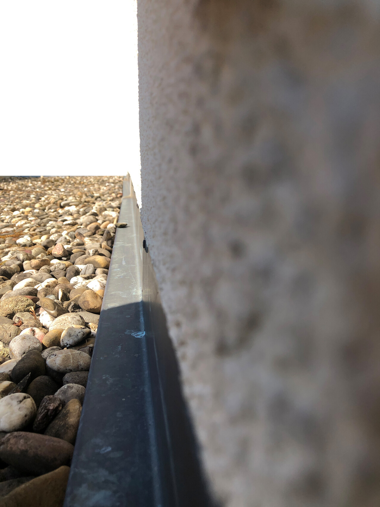 Close-up side view of the building: gravel on the left, grey metal edge at the foundation, bright sky.