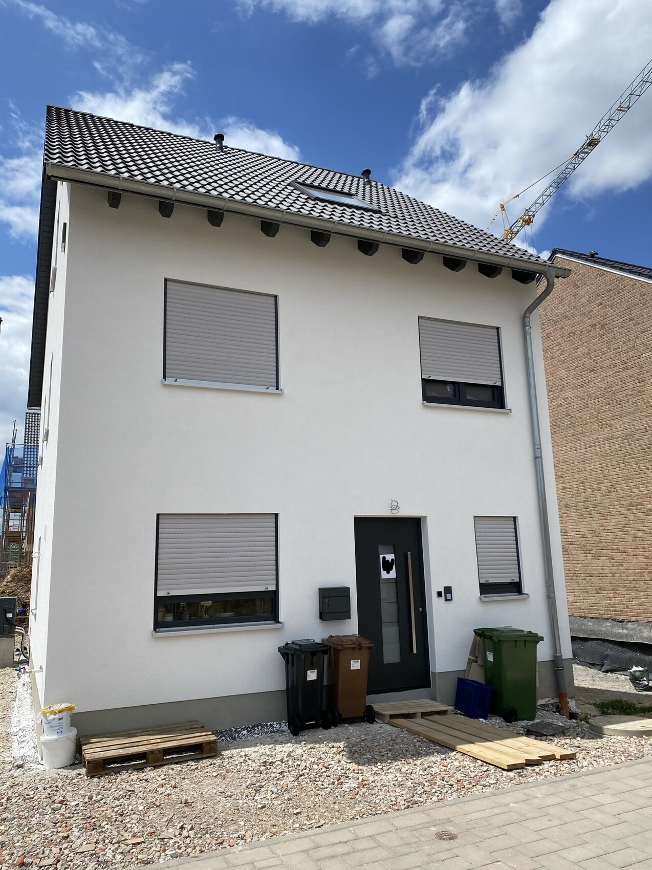 White two-story house under construction with roller shutters; garbage bins, pallets in front of the door.