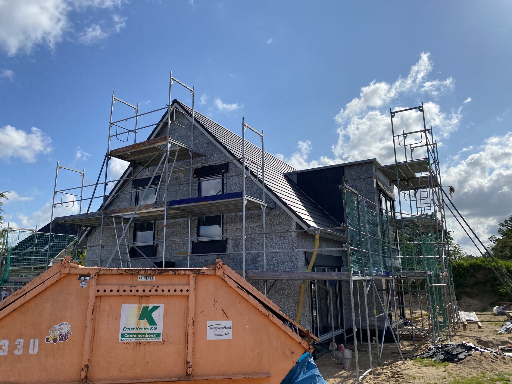 Two-story house with scaffolding, orange site container in front, blue sky.