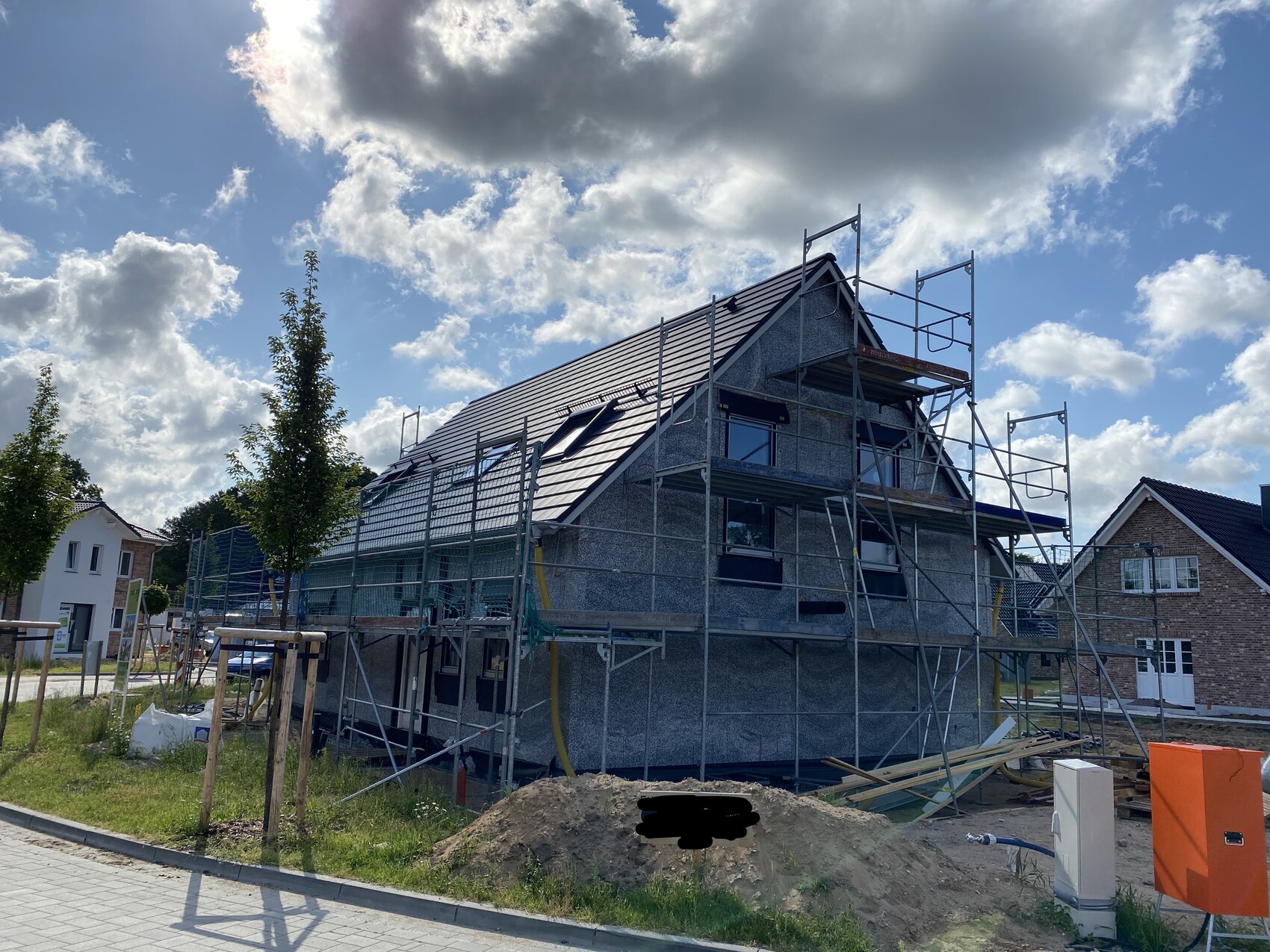 Single-family house under construction with scaffolding around the roof, other houses nearby and cloudy sky.