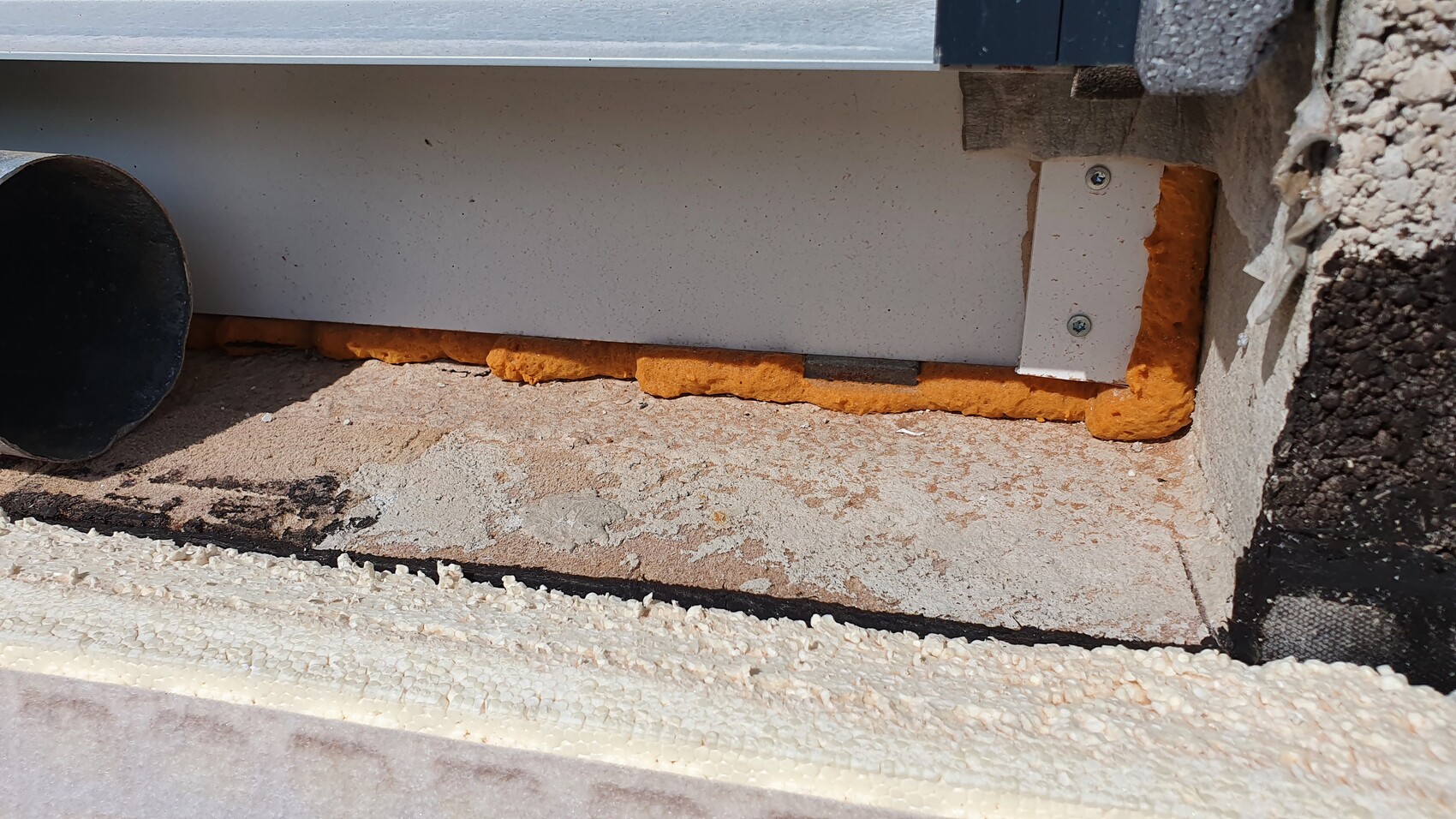 Außenwand-Fugenbereich mit orange Dichtschaum, weiße Verkleidung und schwarzes Rohr.