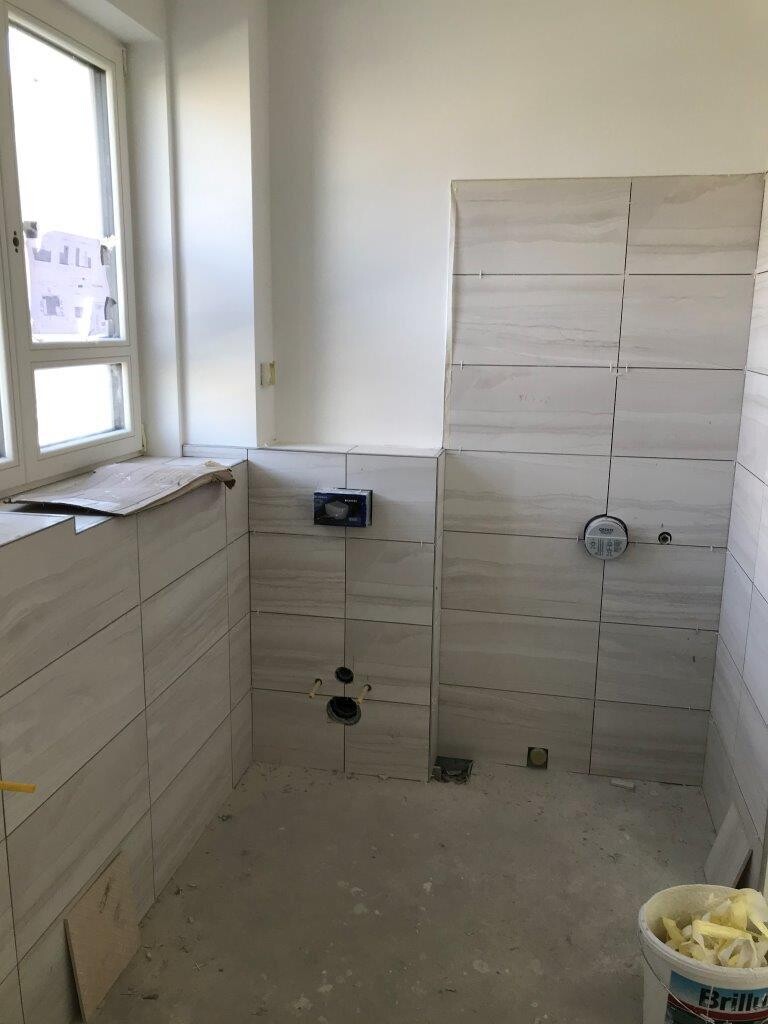 Bathroom under construction, light gray tiles, window on the left, visible installations, bucket on the right.