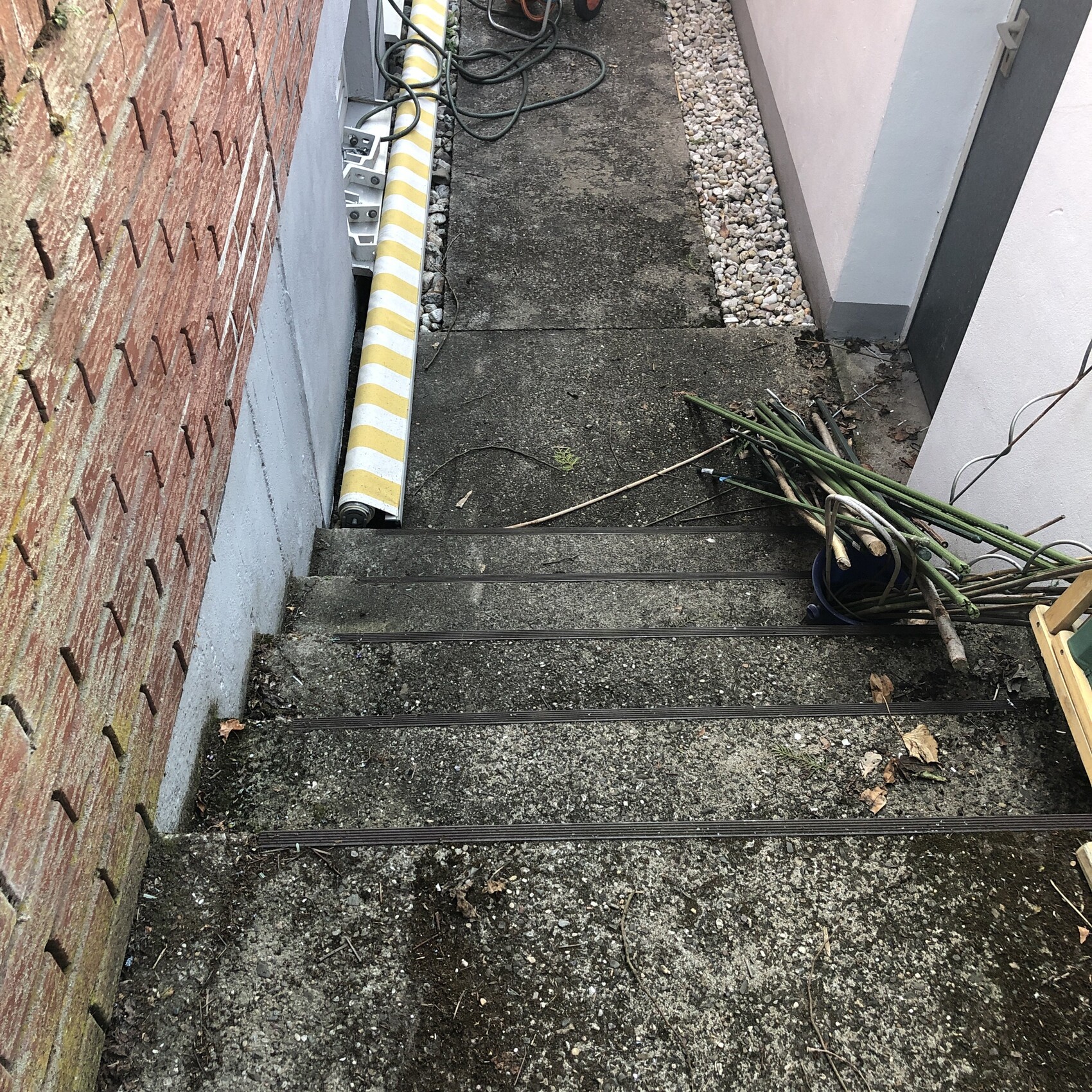 Außenstufen aus Beton mit Kies, linke Ziegelwand, gelbe Schutzrolle, Schläuche und Werkzeuge rechts.