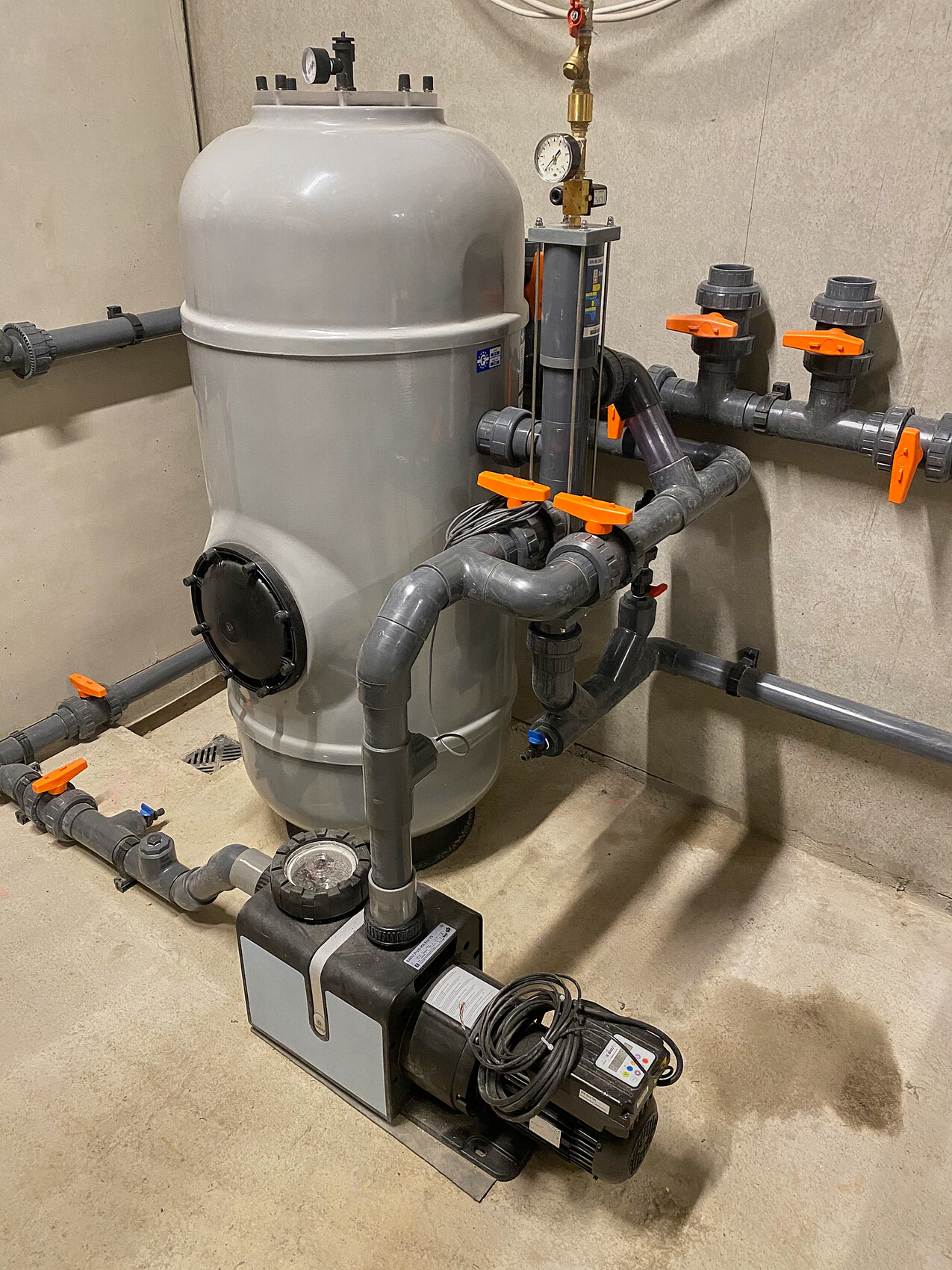 Large gray filter tank with pump, valves, and piping in the technical room