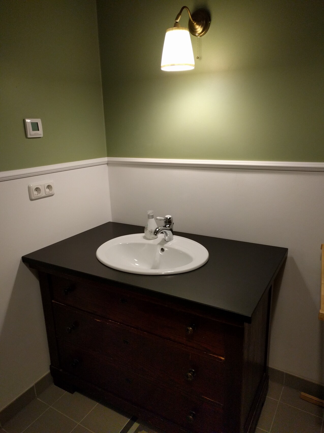White ceramic sink on dark wood base, green wall and wall lamp above.