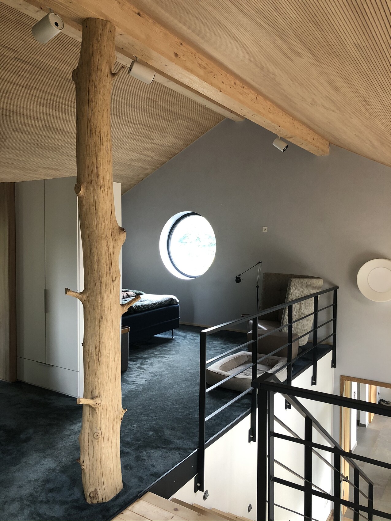 Loft with tree trunk as support, round window, bed and reading chair.