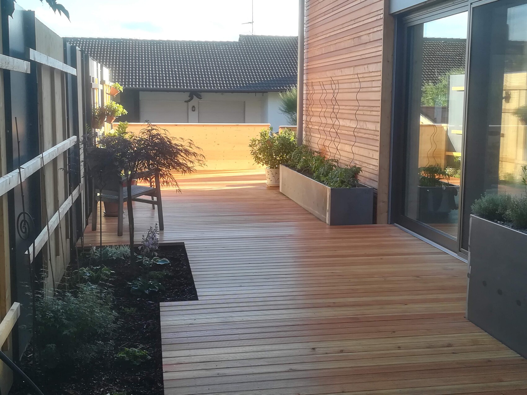 Well-maintained wooden terrace with planter boxes, fence on the left, house wall and glass door on the right.