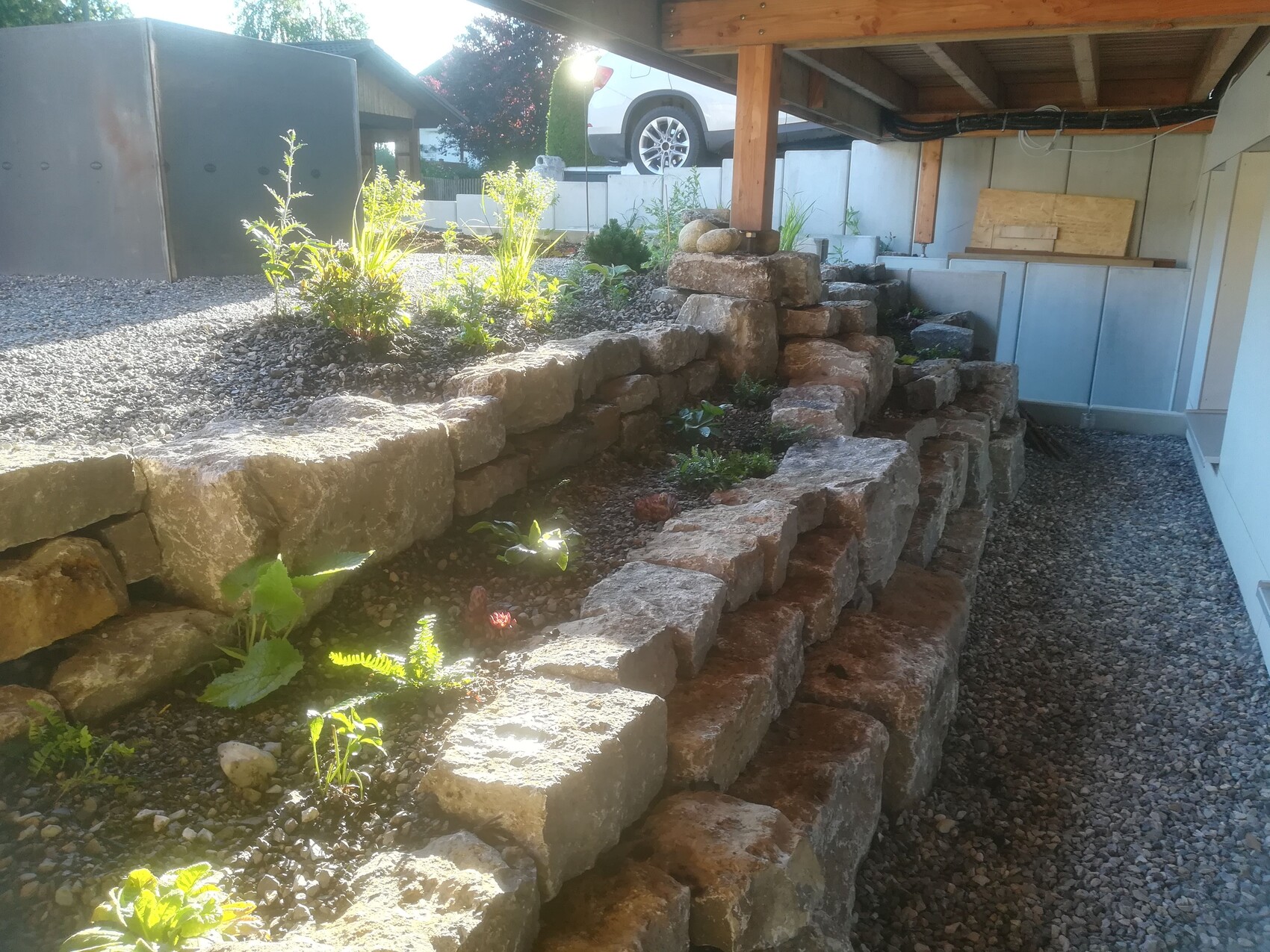 Multi-level natural stone wall made of large blocks next to gravel path, carport in the background