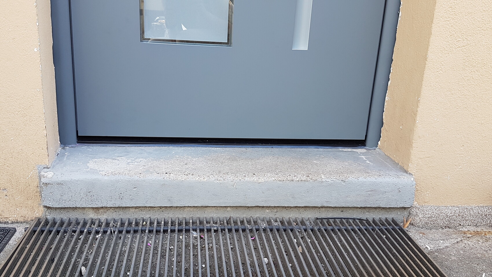 Außenansicht einer blaugrauen Tür mit Fenster; davor eine Betonstufe und ein Metallgitter.