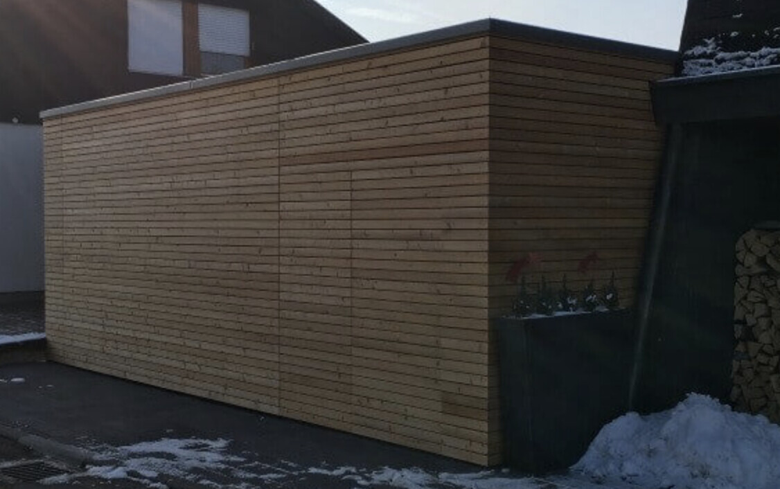 Exterior wall made of horizontal wooden slats; on the right, a planter with red flowers, snow.