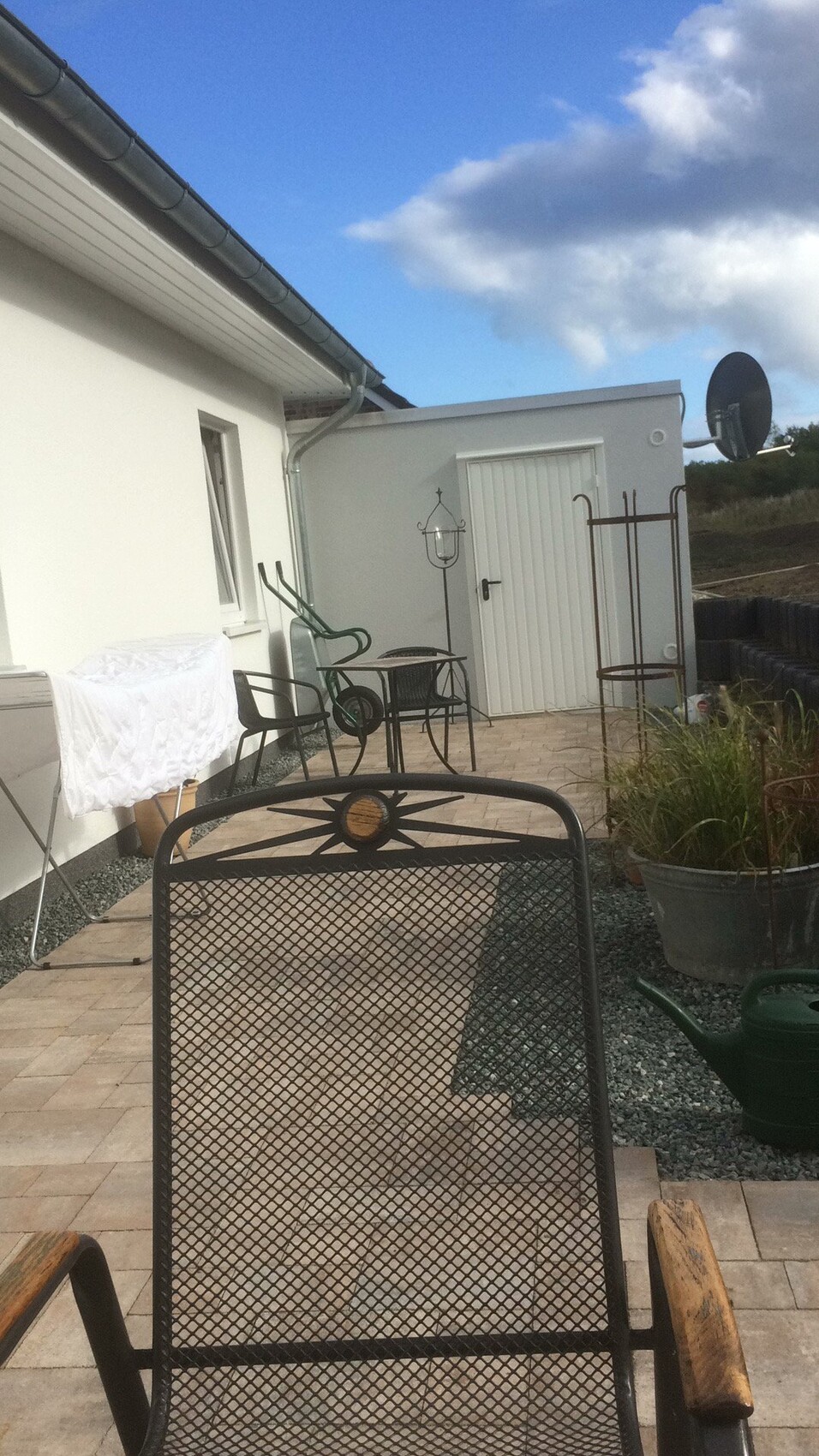 Terrace with metal chair in the foreground, garden furniture and drying rack in front of a white house.