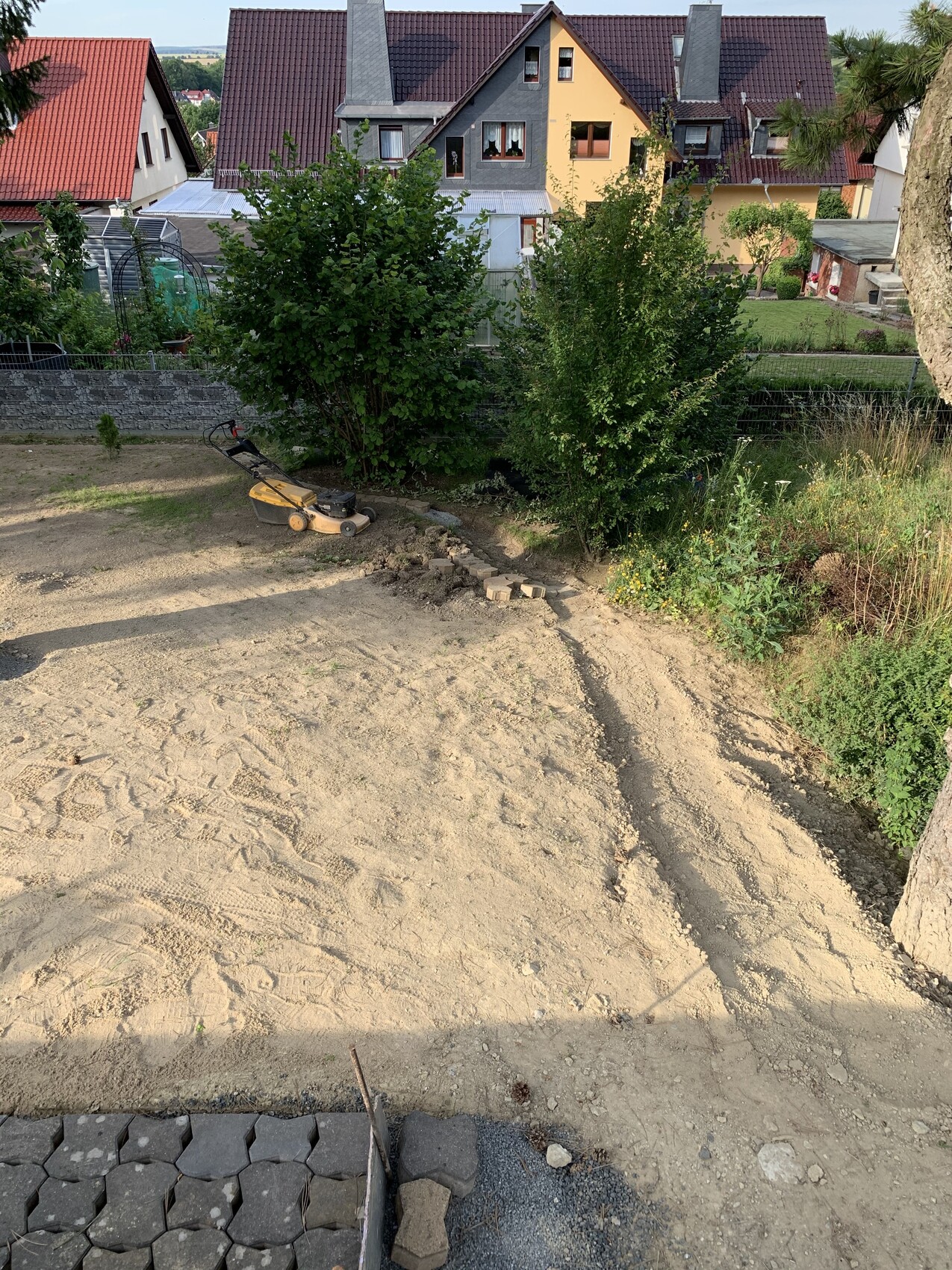 Garden construction site: sandy soil, lawn mower, shrubs, and paving stones with houses in the background.