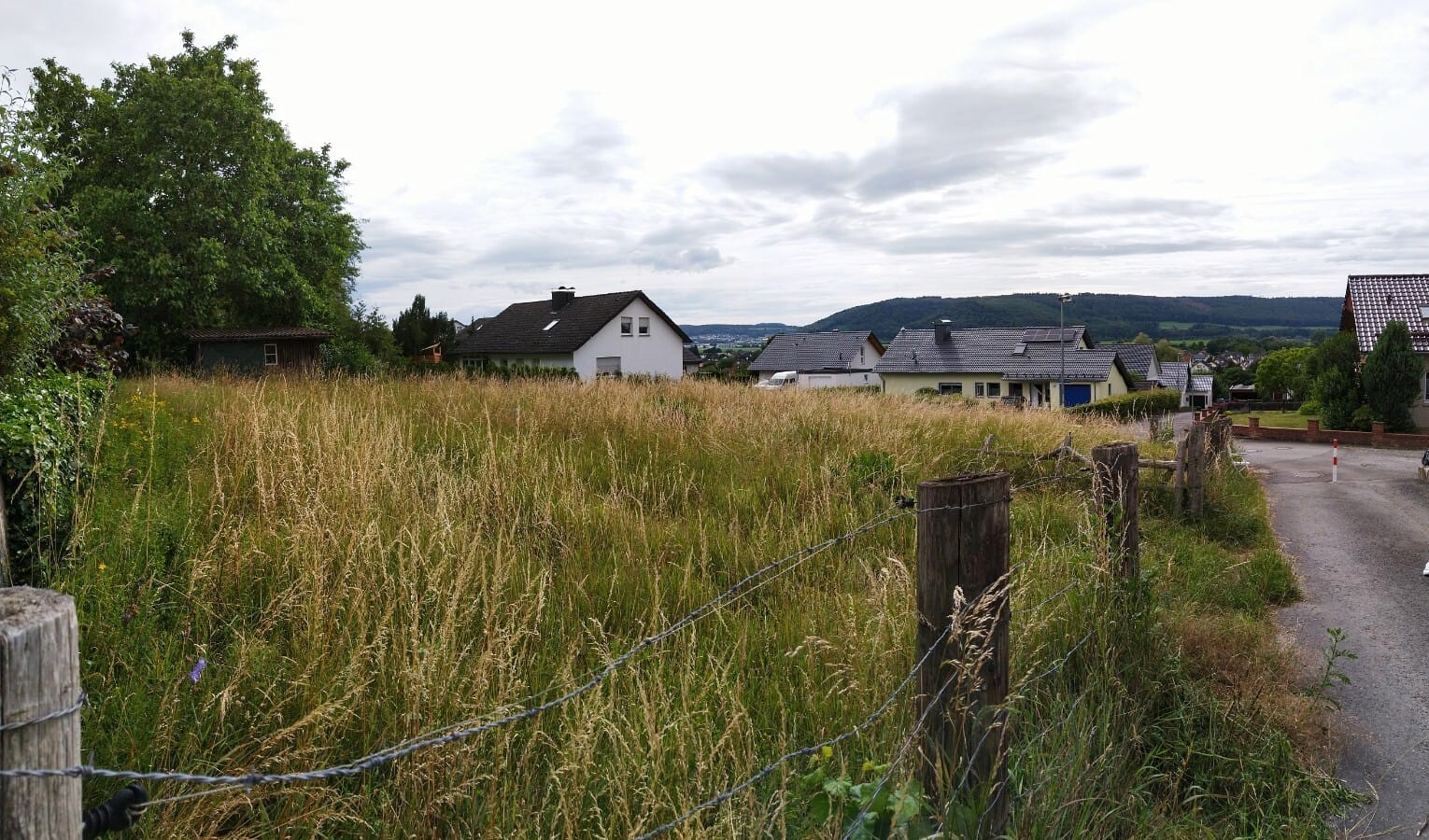 Wide grassy field by the roadside, fence posts with wire, houses and hills in the background.