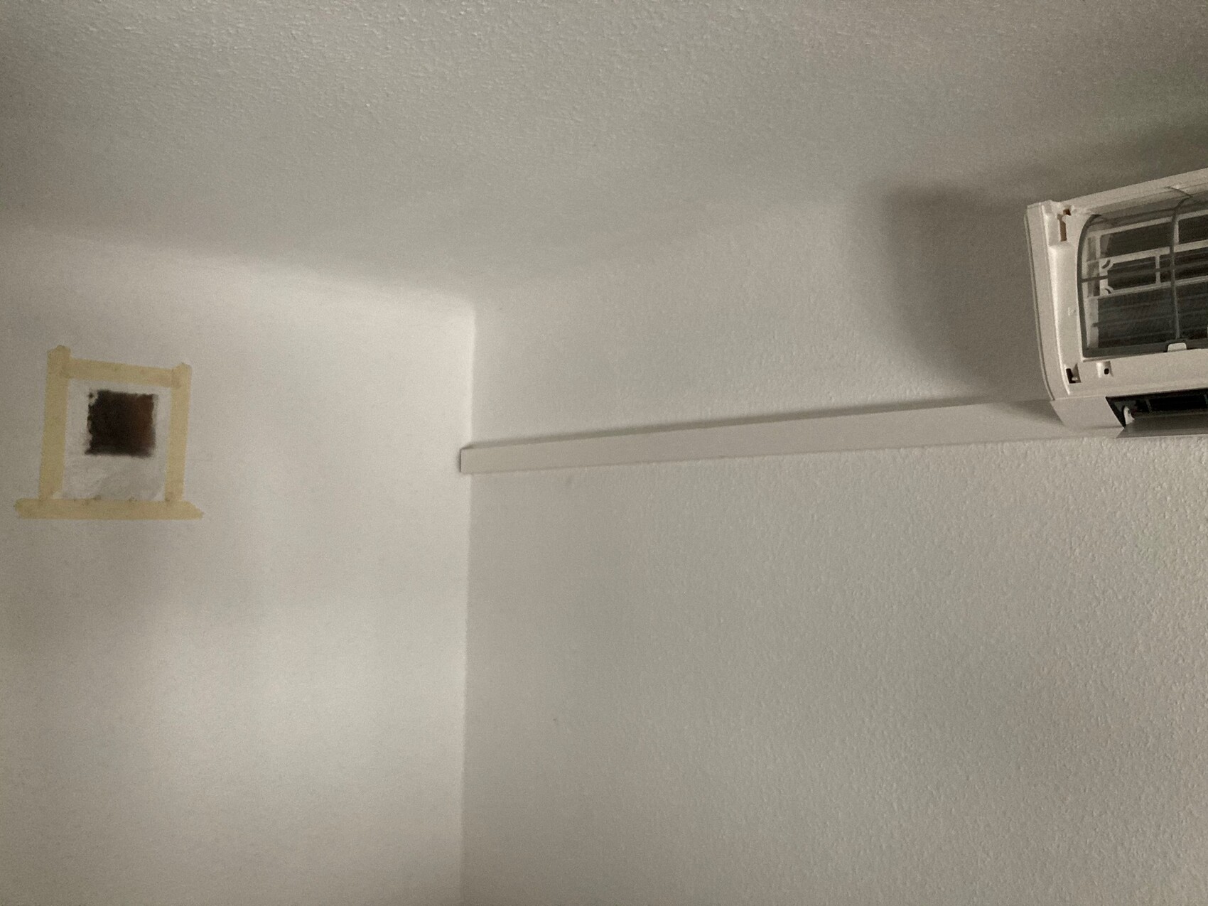 White interior wall with air conditioning unit mounted on the right; brown square behind painter’s tape on the left.